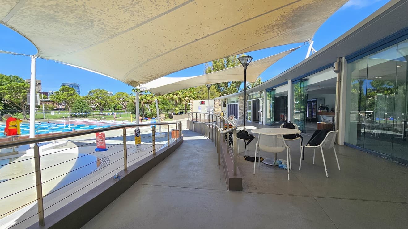 To the right is the cafe enclosed with glass. Outside, movable tables and chairs, equipped with belongings are displayed. A ramp entry, equipped with handrails, leads to the pool area. To the left are the Splash Pool and the 50m Lap Pool, equipped with floating ropes. White shade cloths are placed overhead, providing shade to the seating area. Pool guards and a free-standing banner are visible on the pool deck. Trees surround the area.