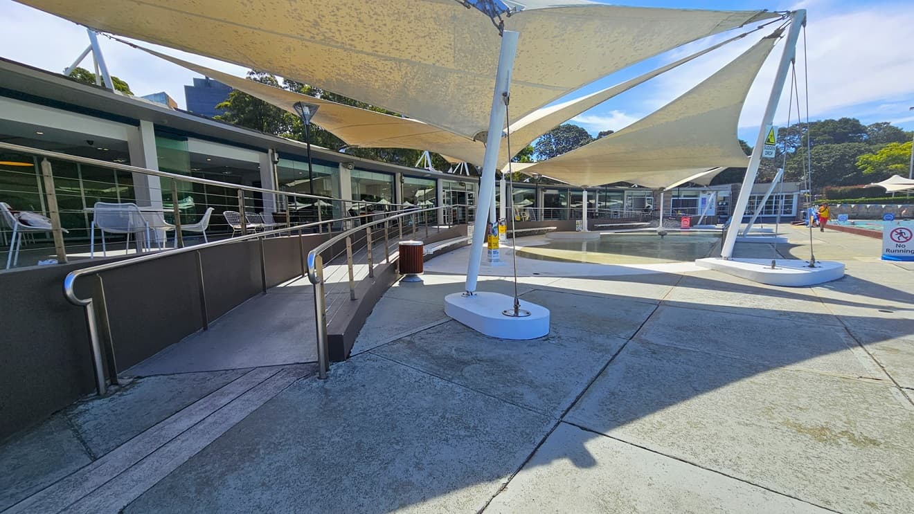 To the left is the cafe enclosed with glass. Outside, movable tables and chairs are displayed. A ramp entry, equipped with handrails, leads to the pool area. To the right are the Splash Pool and the 50m Lap Pool, equipped with floating ropes. White shade cloths are placed overhead, providing shade to the seating area. Pool guards and a free-standing banner are visible on the pool deck. Trees surround the area.