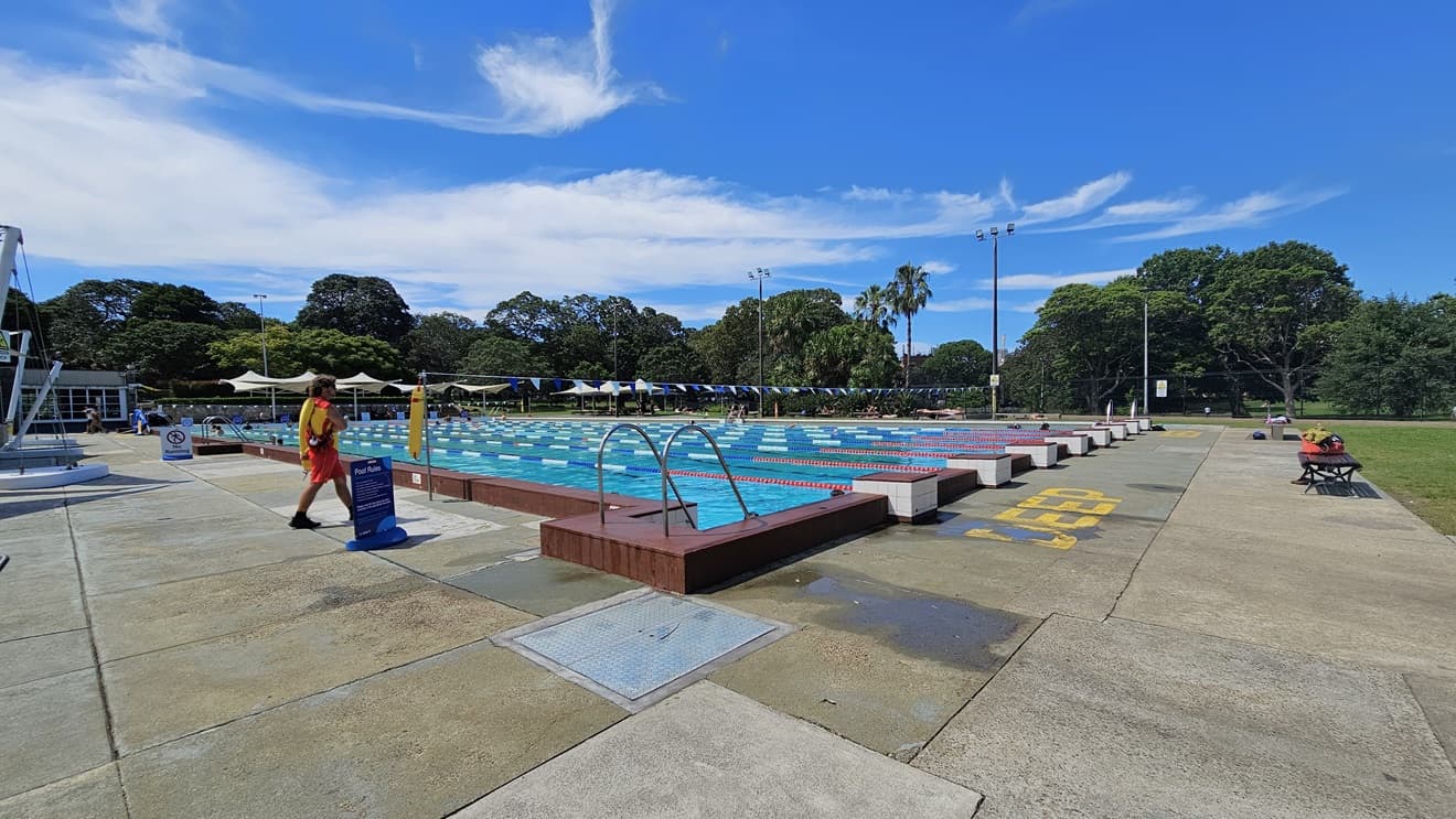 The 50m Lap Pool is equipped with floating ropes and step entry points with handrails. Bench seating with belongings is placed on the right near the grass. The concrete floor has yellow markings indicating the pool's deep water level. Free-standing banners and safety warnings are on the pool deck. A string of blue buntings runs across the pool. Further ahead, umbrella shades are visible, and a pool staff member is present.