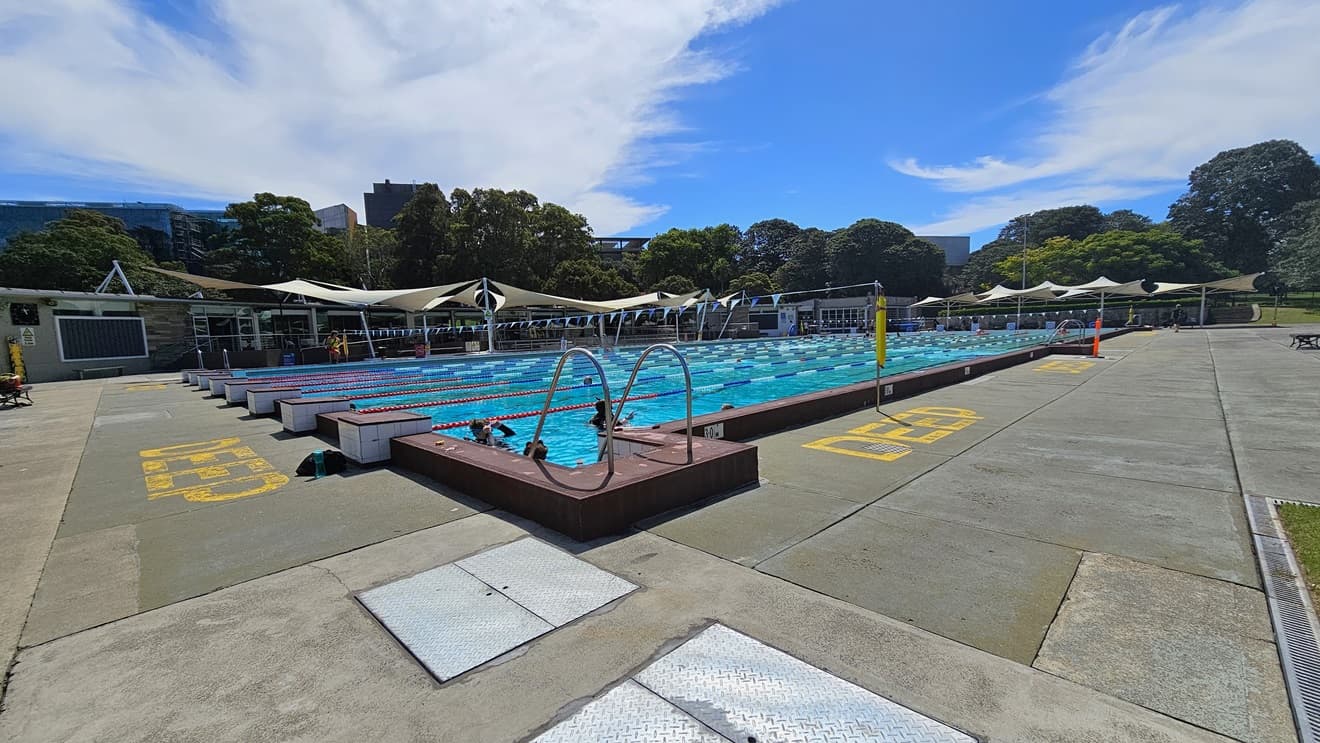 The 50m Lap Pool features floating ropes and step entry points with handrails. The concrete floor is marked with yellow indications for the deep water level. Free-standing banners and safety warnings are present on the pool deck. A string of blue buntings runs across the pool. Further ahead, umbrella shades are visible on the left and far right.