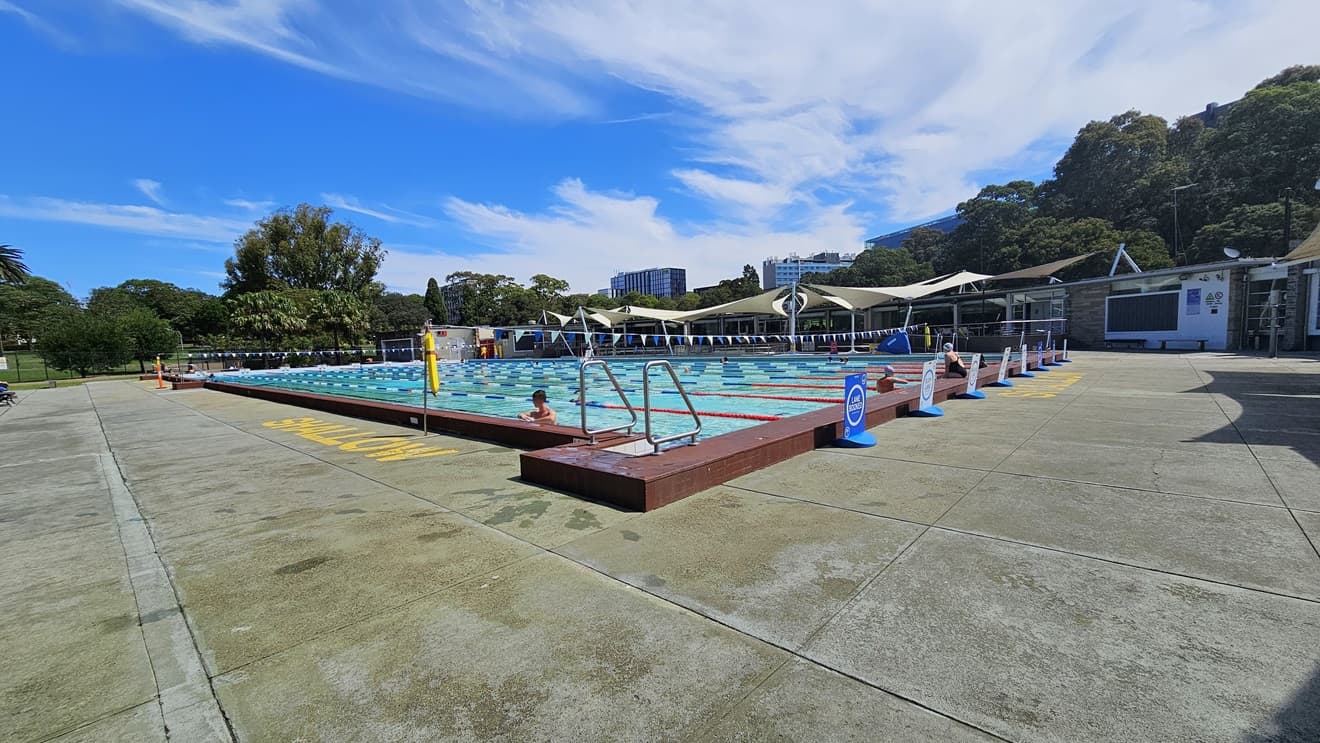 The 50m Lap Pool is equipped with floating ropes and step entry points with handrails. The concrete floor is marked with yellow indications for the shallow water level. Free-standing banners are positioned on the pool deck. A string of blue buntings runs across the pool. Further ahead, umbrella shades are visible on the right, adjacent to a brick building. Tall trees surround the area.