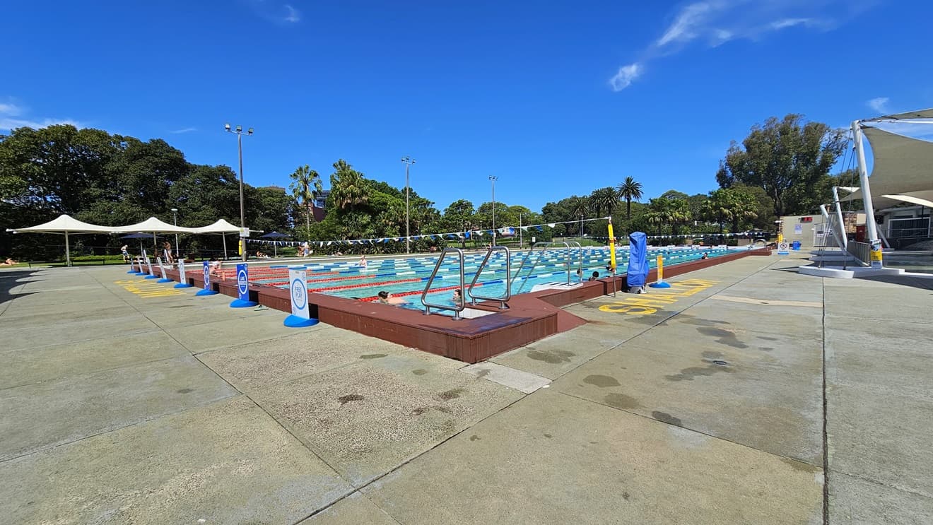 The 50m Lap Pool has floating ropes and step entry points with handrails. The concrete floor is marked with yellow indications for the shallow water level. On the left, free-standing banners are positioned on the pool deck. A string of blue buntings runs across the pool. To the left, umbrella shades are visible, and shade cloths are on the right. A hoist is positioned near the pool. Tall trees surround the area.