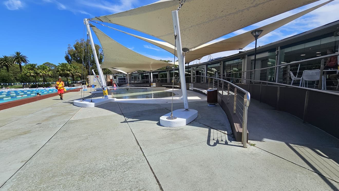 To the right is the cafe enclosed with glass. Outside, movable tables and chairs are displayed. A ramp entry, equipped with handrails, leads to the pool area. To the left is the 50m Lap Pool, equipped with floating ropes. White shade cloths are placed overhead, providing shade to the seating area. Pool guards and a free-standing banner are visible on the pool deck. Trees surround the area.