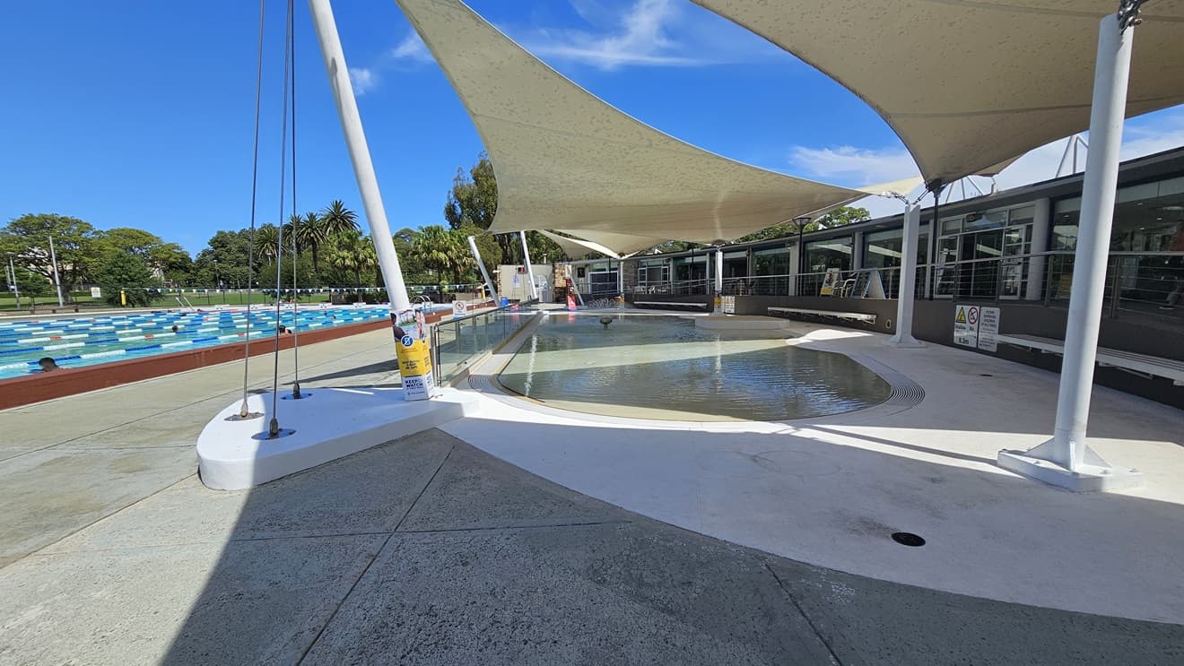 To the right is a ramp entry, equipped with handrails, leading to the pool area. In the middle is the Kid's Splash Pool with a beach entry and a small water fountain. To the left is the 50m Lap Pool, equipped with floating ropes. White shade cloths are placed overhead, providing shade to the seating area. Free-standing banners and safety signs are visible. Trees surround the area.