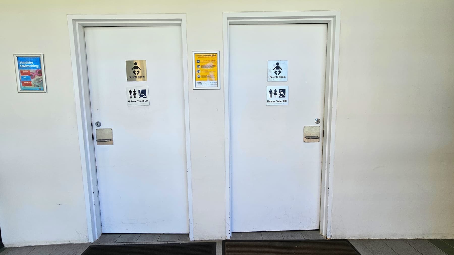 wo white doors lead to the accessible unisex bathroom and change rooms. A poster is affixed to the middle and to the right. Non-slippery mats are placed on the doorsteps. The wall is white, and the floor has grey tiles.
