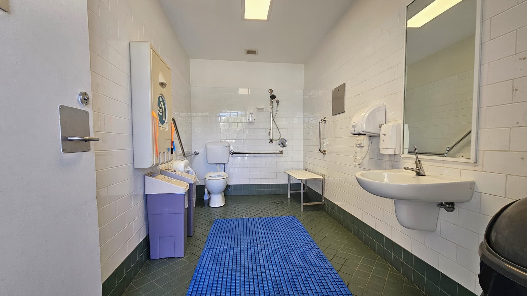 Inside the accessible bathroom, the toilet bowl equipped with handrails is in the left corner. Adjacent to it is the shower, with hand rails and a foldable bench affixed to the middle wall. To the right is a washbasin, mirrors, along with a liquid soap and tissue dispenser. A foldable change table is mounted to the left wall, and below it are two rubbish bins. The grey floor is equipped with a blue non-slippery mat, and the ceiling and wall are white.