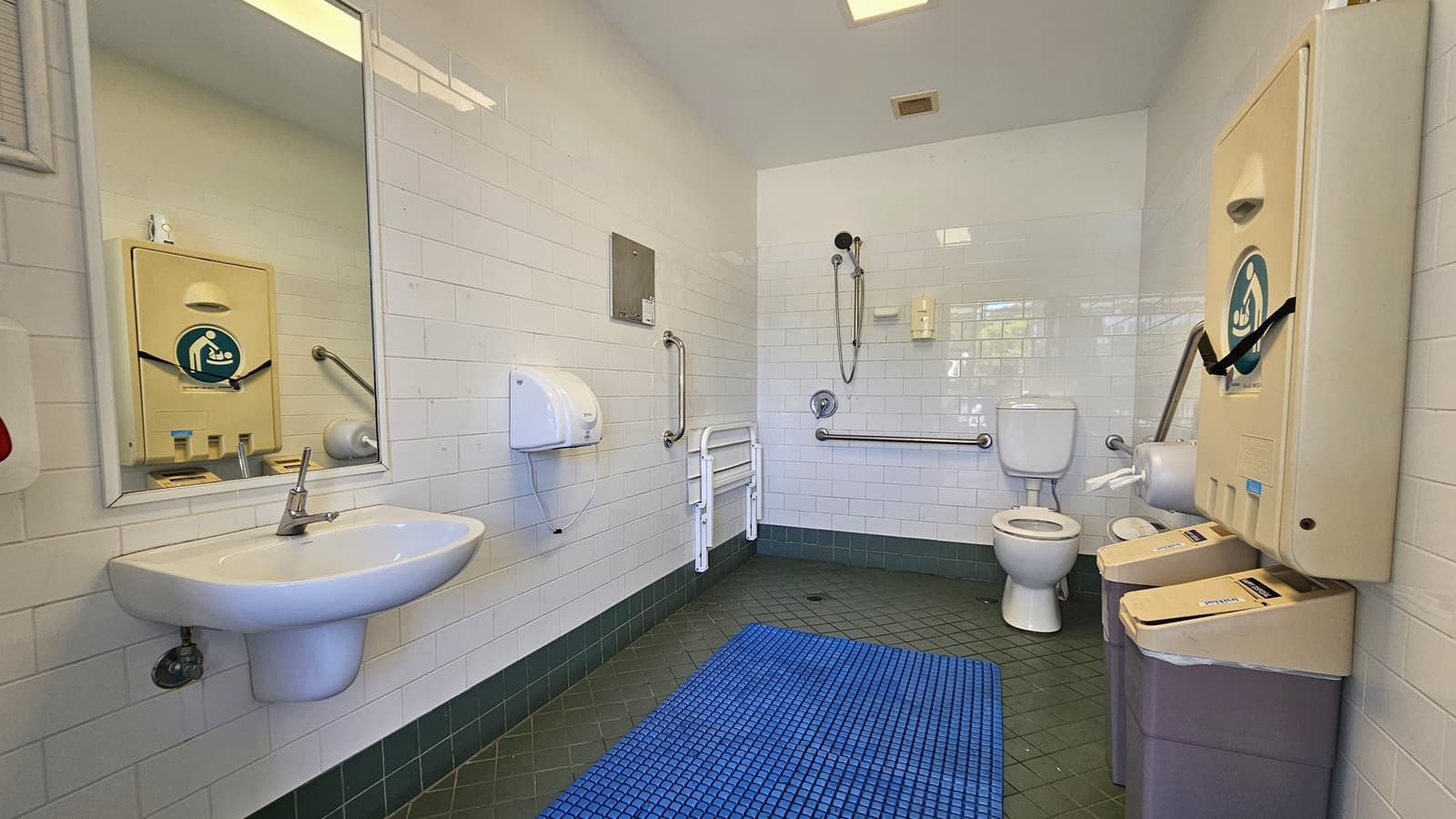 Inside the accessible bathroom, the toilet bowl equipped with handrails is in the right corner. Adjacent to it is the shower, with hand rails and a foldable bench affixed to the left wall. To the left is a washbasin, mirrors, along with a liquid soap and tissue dispenser. A foldable change table is mounted to the right wall, and below it are two rubbish bins. The grey floor is equipped with a blue non-slippery mat, and the ceiling and wall are white.