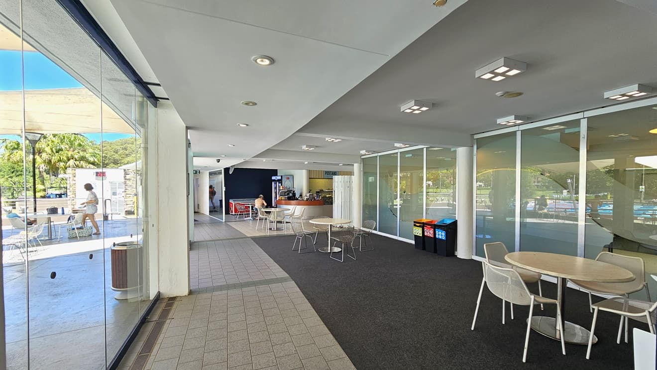 Movable tables and chairs are arranged to the right of the glass walls and in front of the cafe counter. The cafe features a timber and silver desk. An ice cream freezer and a fridge for cold drinks are positioned to the left and right of the counter. The floor is a combination of grey tiles and grey carpet. The ceiling is white with square and round fluorescent lights.