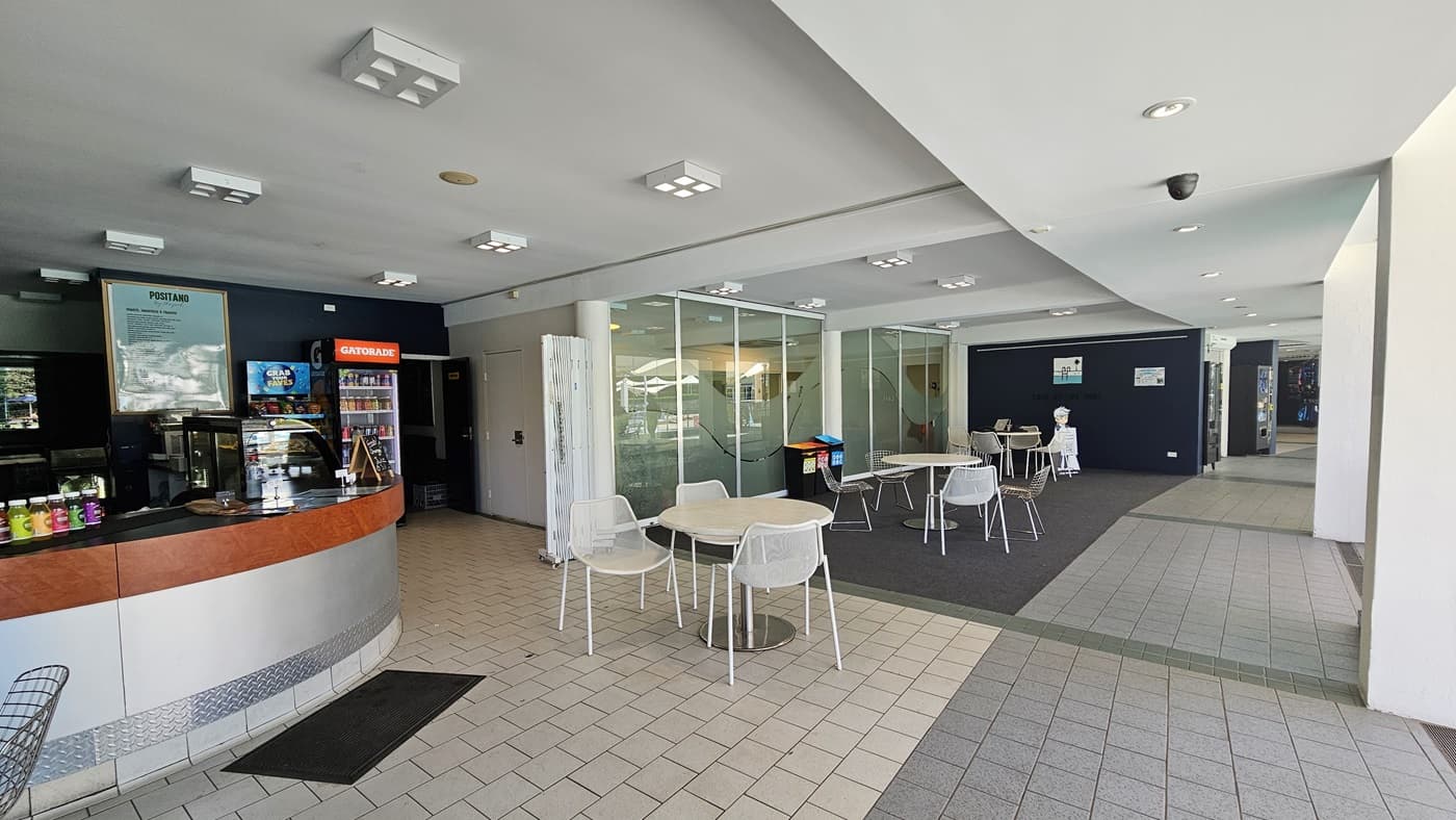Movable tables and chairs are arranged to the right of the glass walls and in front of the cafe counter. The cafe features a timber and silver desk. Food items and food racks are on top of the counter. A fridge displaying cold drinks stands on the right of the counter. The floor is a combination of grey tiles and grey carpet. The ceiling is white with square and round fluorescent lights. A black non-slip mat is also in place.