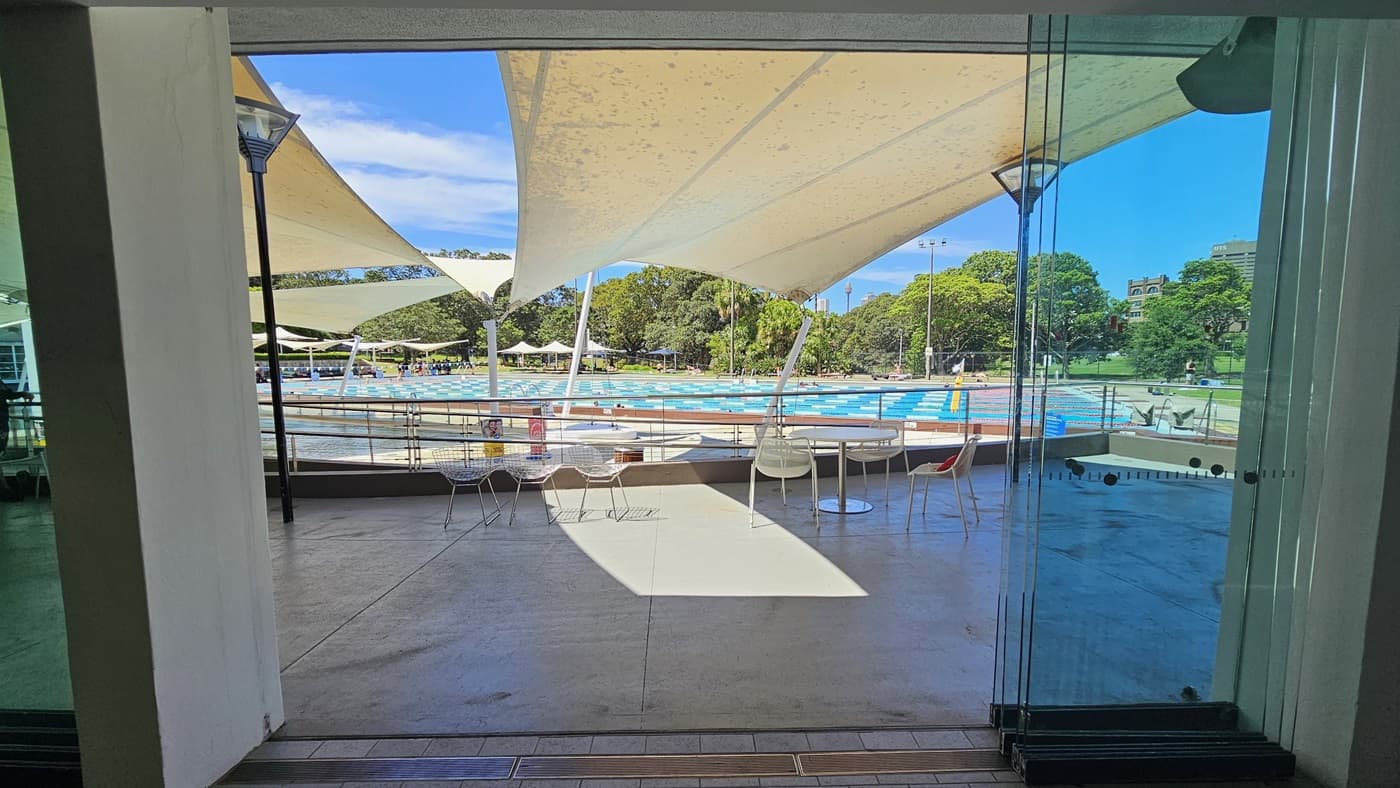 Movable tables and chairs are arranged in front of the cafe, near the pool deck. Multiple white cloth shades are placed overhead. Adjacent is the ramp entry equipped with handrails that leads to the pool. Lamp posts are also in place. Further behind is the 50m lap pool equipped with floating ropes. Umbrella shades and trees are visible further behind.