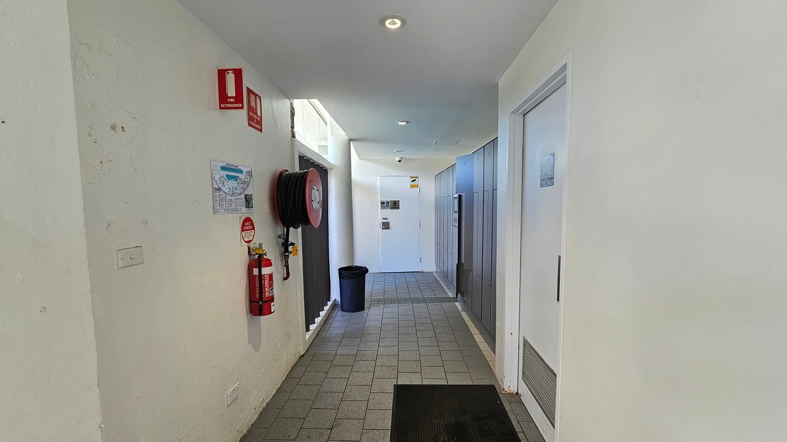 To the left is a fire hose reel and fire extinguisher mounted on the wall, equipped with signage. A poster bearing the site map is also affixed to the wall. To the right and further in are the doors leading to the accessible bathroom. A rubbish bin and a non-slip mat is also present. The floor has grey tiles, and the ceiling and wall are white with LED lights.