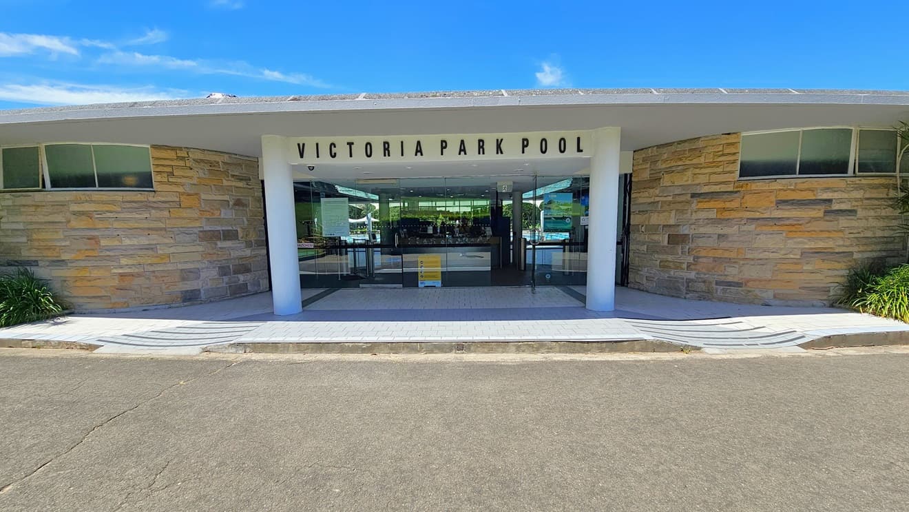 The facility entrance showcases a brown concrete facade with a portico displaying the facility's name in bold letters. Step-free access is available on both sides. Landscaped plants adorn the left and right areas. The entrance comprises a manual glass door with posters and a freestanding sign in the middle. The floor transitions from concrete to white bricks with grey accents.