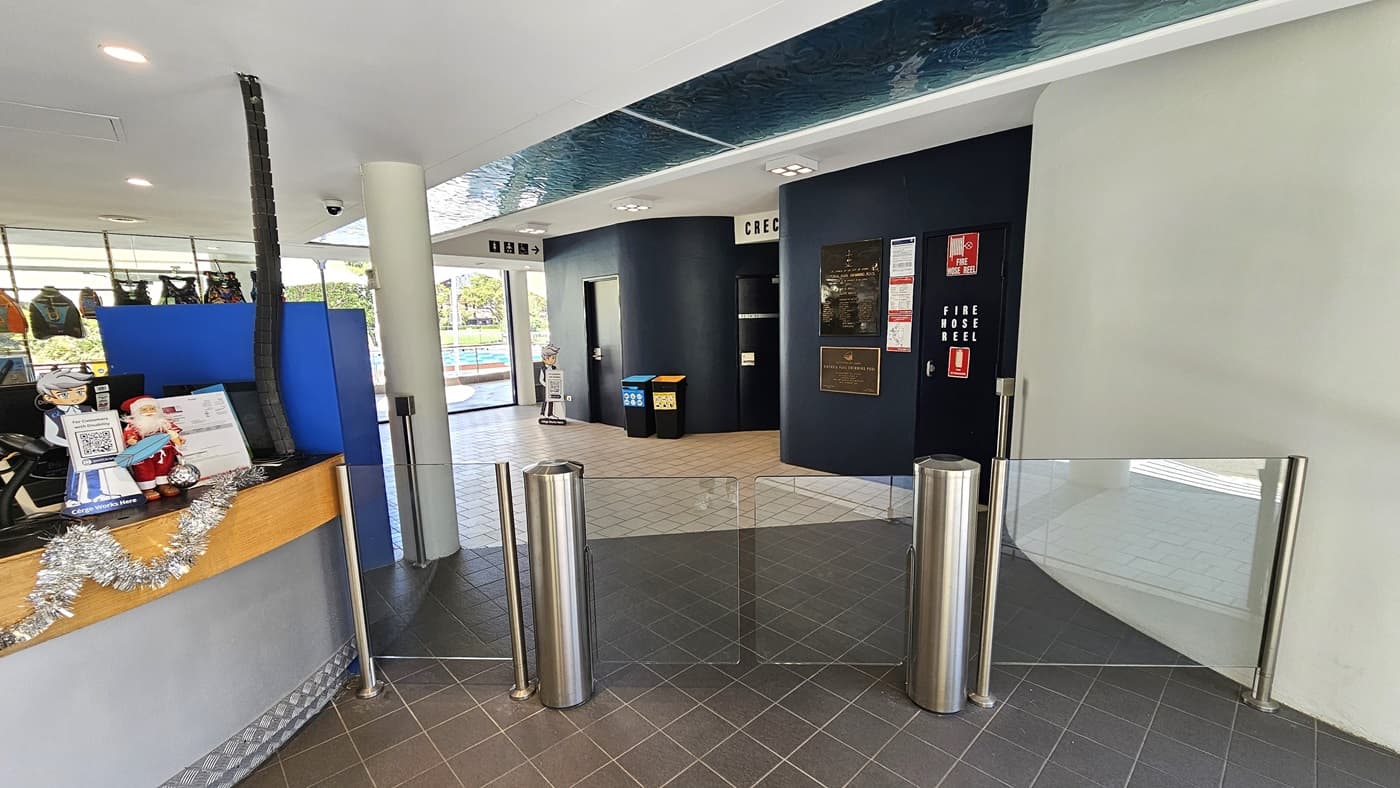 The reception desk, featuring a circular design with silver and timber elements, is centrally located. The counter is decorated with Christmas garland, card swipe machines, computers, and brochures. A turnstile glass gate is positioned to the right. Inside, a black wall displays posters and directional signage, with rubbish bins in place. The floor has grey tiles, and the ceiling is white with an aqua accent. Behind the desk, retail items are on display.