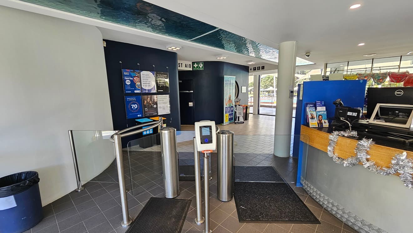 The centrally located reception desk has a circular design with silver and timber elements. The counter is adorned with Christmas garlands, computers, and brochures. To the left, a turnstile glass gate is equipped with a swiping machine and a non-slippery mat. Inside, a black wall displays posters and directional signage, with rubbish bins in place. The floor is covered in grey tiles, and the ceiling is white with an aqua accent. Behind the desk, retail items are on display.