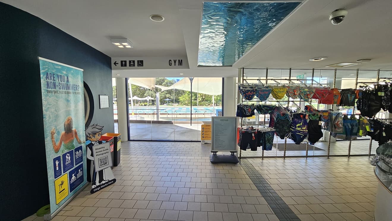 To the right is a retail area displaying swimwear. To the left, there's a large retractable banner, and two rubbish bins are positioned against the black wall. Directional signages are affixed to the upper left near the ceiling. Behind, a glass wall provides a view of the outdoor pool. The floor is covered in grey tiles, and the ceiling is white with an aqua accent.