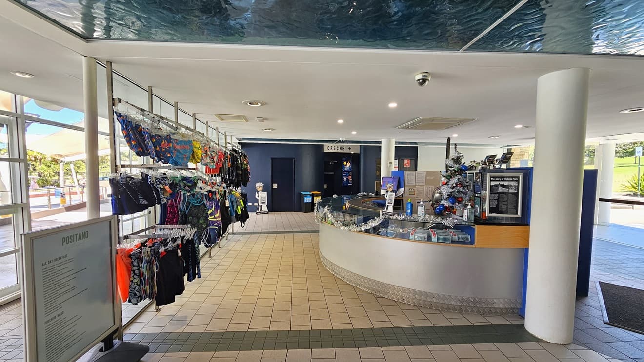 The reception desk, designed with a circular pattern using silver and timber elements, is located on the right. The counter is adorned with Christmas garland, computers, and brochures. On the left, swimwear is displayed for sale, along with a banner for the Positano cafe. The floor has brown tiles, and the ceiling is white with an aqua accent.