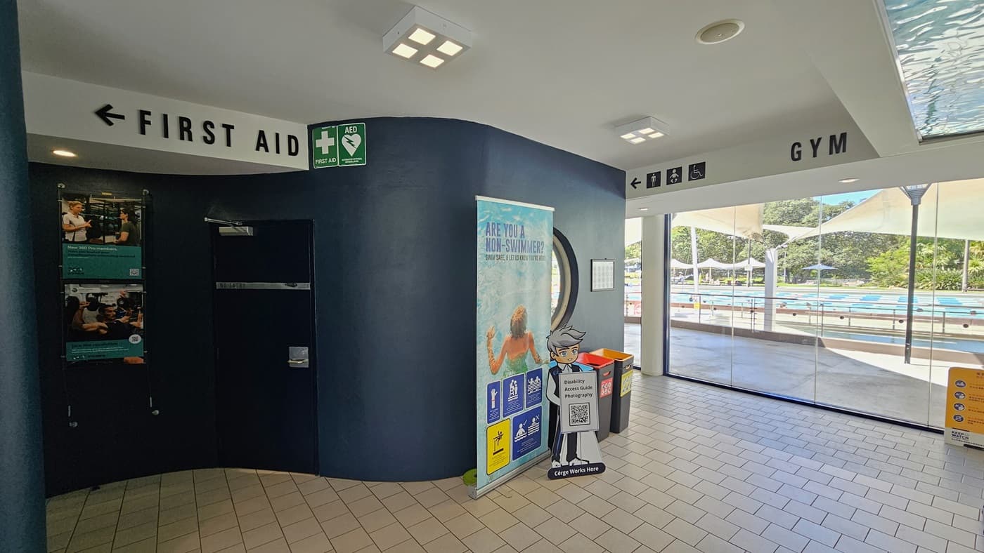 To the left, there's a black wall with posters and directional signages. A "First Aid" sign is affixed to the upper left. A large retractable banner is against the black wall with rubbish bins beside it. To the right is a glass wall and a yellow sign. Behind the glass wall is the outdoor aquatic facility. The floor is covered in grey tiles, and the ceiling is white with an aqua accent.