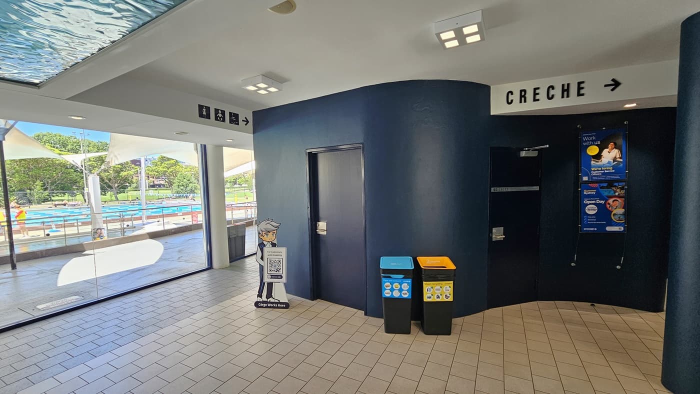 To the right, there's a black wall with posters and directional signages. A "Creche" sign is affixed to the upper right, and accessible bathroom signs are affixed to the upper left. Two rubbish bins are placed against the black wall. To the left is a glass wall, behind which is the outdoor aquatic facility. The floor is covered in grey tiles, and the ceiling is white with an aqua accent.