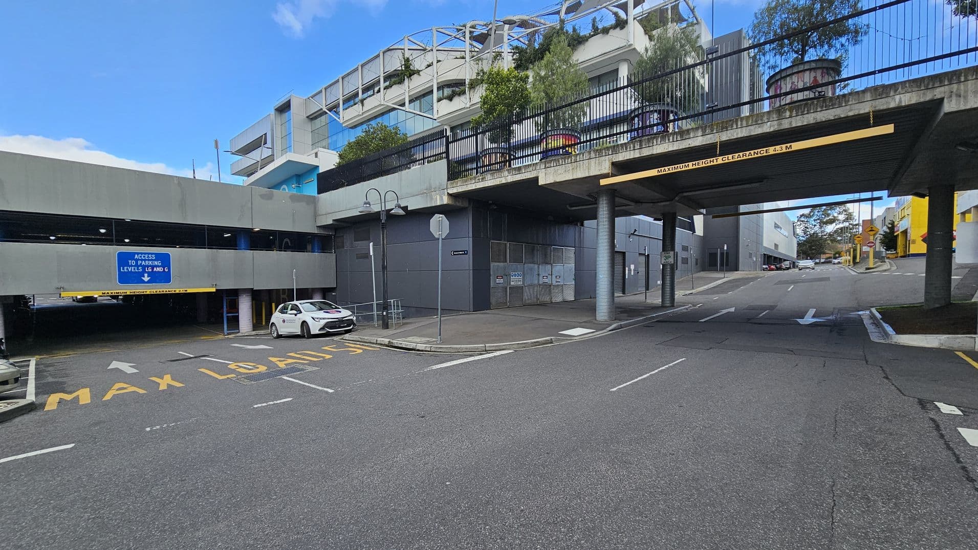 A multi-level car park entrance is on the left, with a blue sign indicating access to parking levels LG and G. A white car is parked nearby. The road has "MAX LOAD 15" painted on it. On the right, a road passes under a bridge with a clearance sign of 4.3 metres. Above the bridge, there are plants and a structure with a metal framework. The background features modern buildings with glass and concrete elements. The road surface is asphalt.