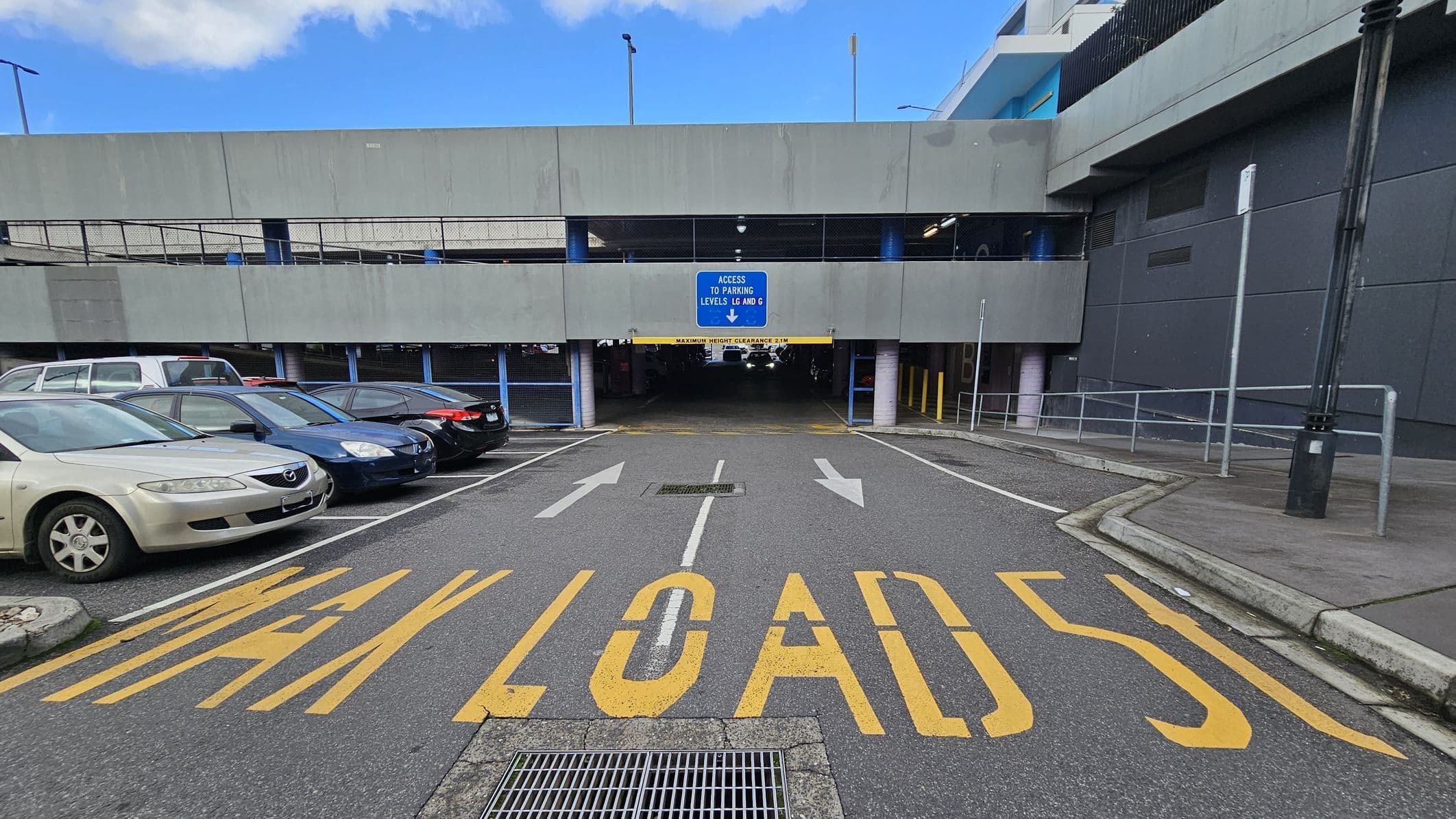 A multi-storey car park entrance with a sign indicating access to parking levels LG and G. The foreground has yellow road markings reading "MAX LOAD 5T" and white arrows pointing towards the entrance. On the left, several parked cars are visible, including a beige sedan. On the right, a pavement with a railing and a tall lamp post. The structure is grey with a blue sign above the entrance. The ground is asphalt, and the ceiling of the entrance is low with a clearance sign.