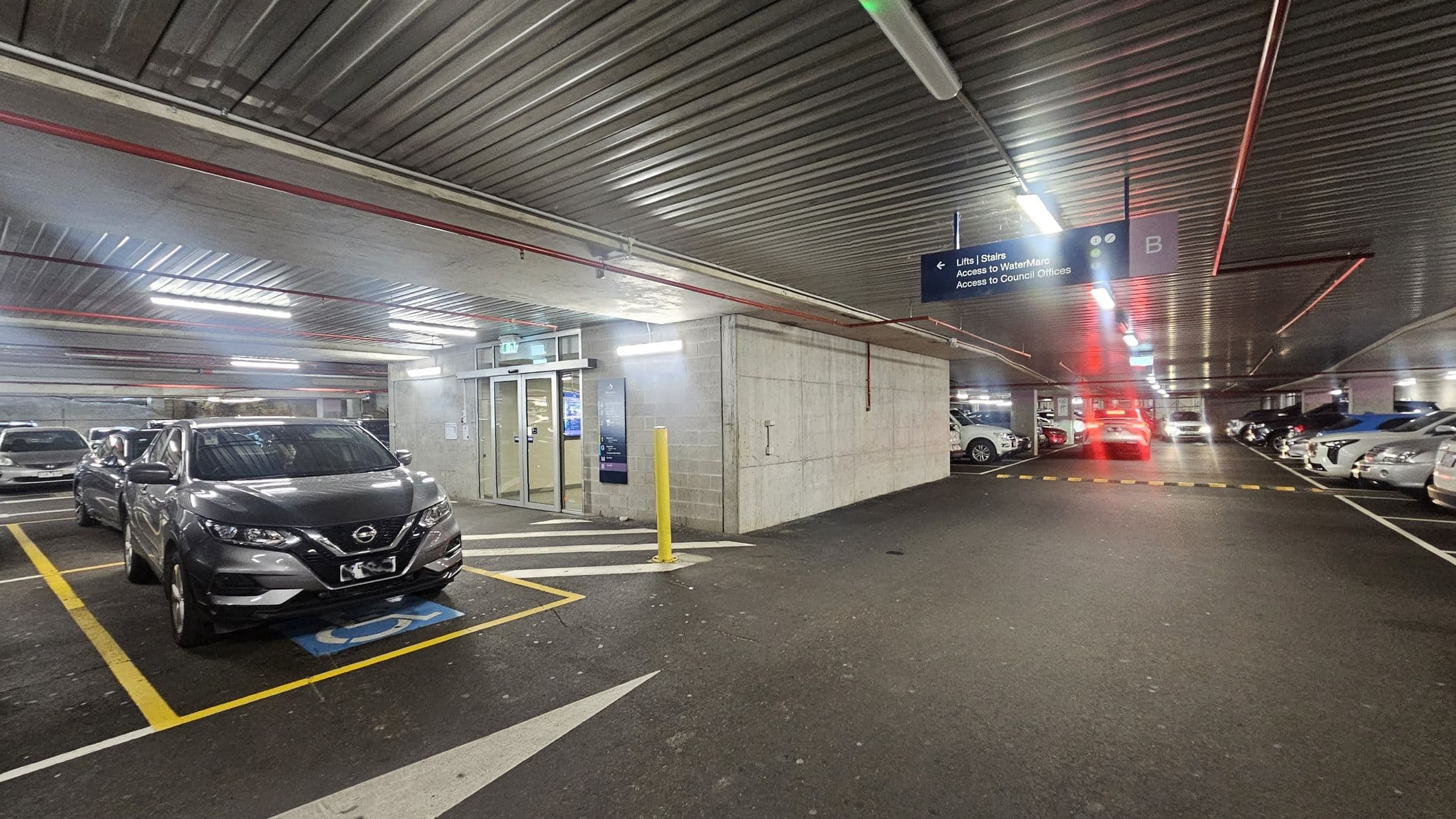 A multi-storey car park with a grey car parked in a designated disabled space on the left. The floor is marked with yellow lines and a blue disabled symbol. In the centre, there is a concrete wall with glass doors leading to lifts and stairs. A yellow bollard is in front of the entrance. On the right, several cars are parked in marked spaces. Overhead, a metal ceiling with fluorescent lights and a sign indicating directions to lifts, WaterMarc, and council offices.