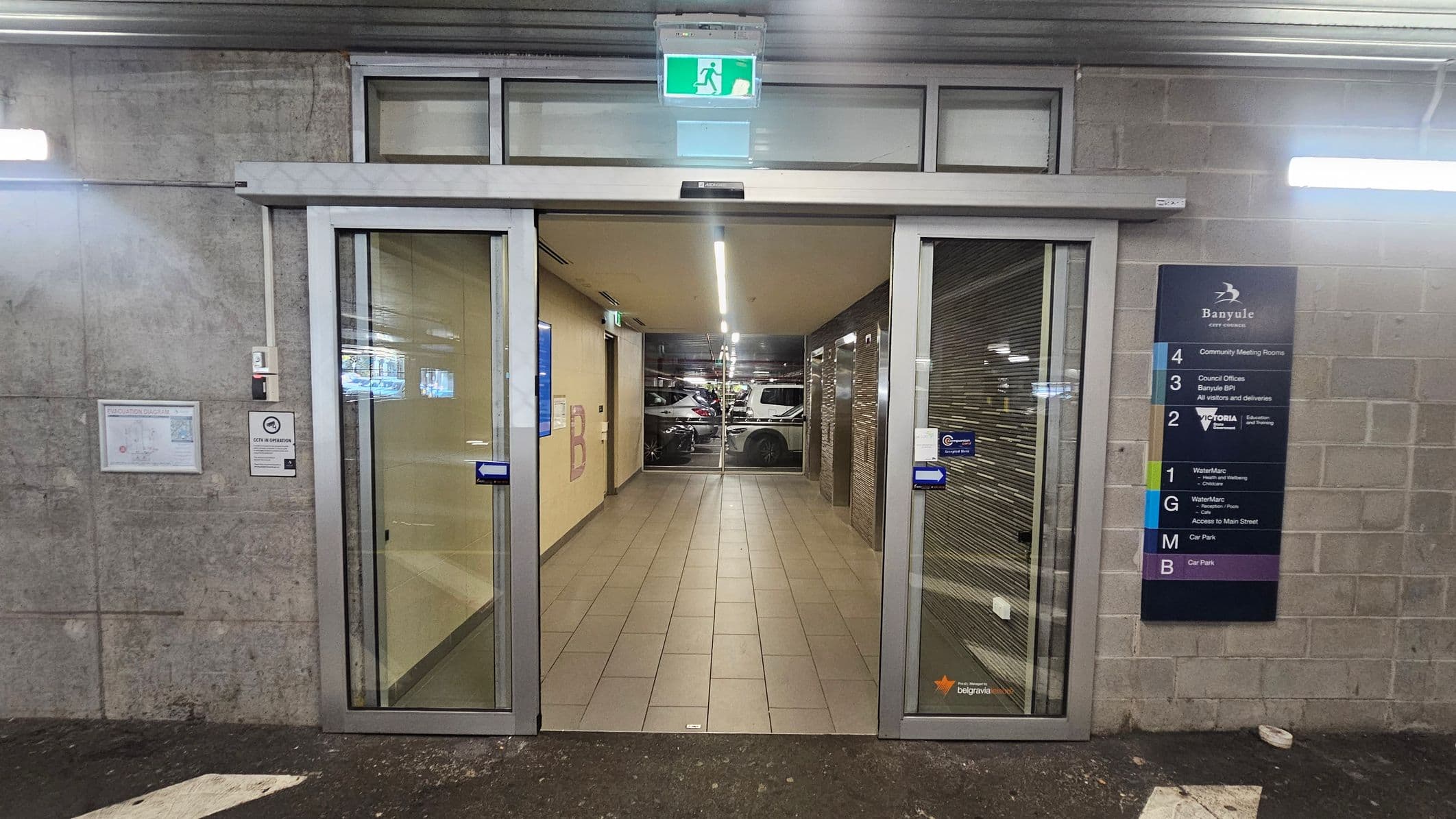Automatic sliding glass doors in the centre, leading to a tiled hallway with a car park visible at the end. On the right, a directory sign lists various floors and facilities, including community meeting rooms and council offices. The left wall has a map and a sign with instructions. The ceiling has a green exit sign above the doors, and the floor outside is dark with a small white section.