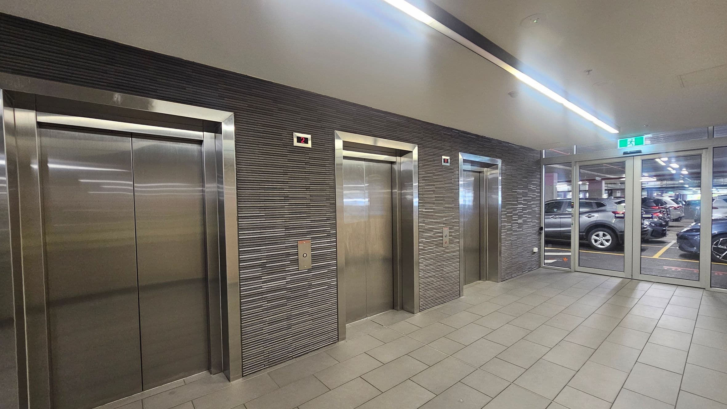 Three stainless steel lift doors are set into a textured grey wall. Above each lift is a digital display showing the number "2". To the right, glass doors lead to a car park with several parked vehicles visible. The floor is tiled in a light grey, and the ceiling features linear fluorescent lighting. An illuminated green exit sign is above the glass doors.