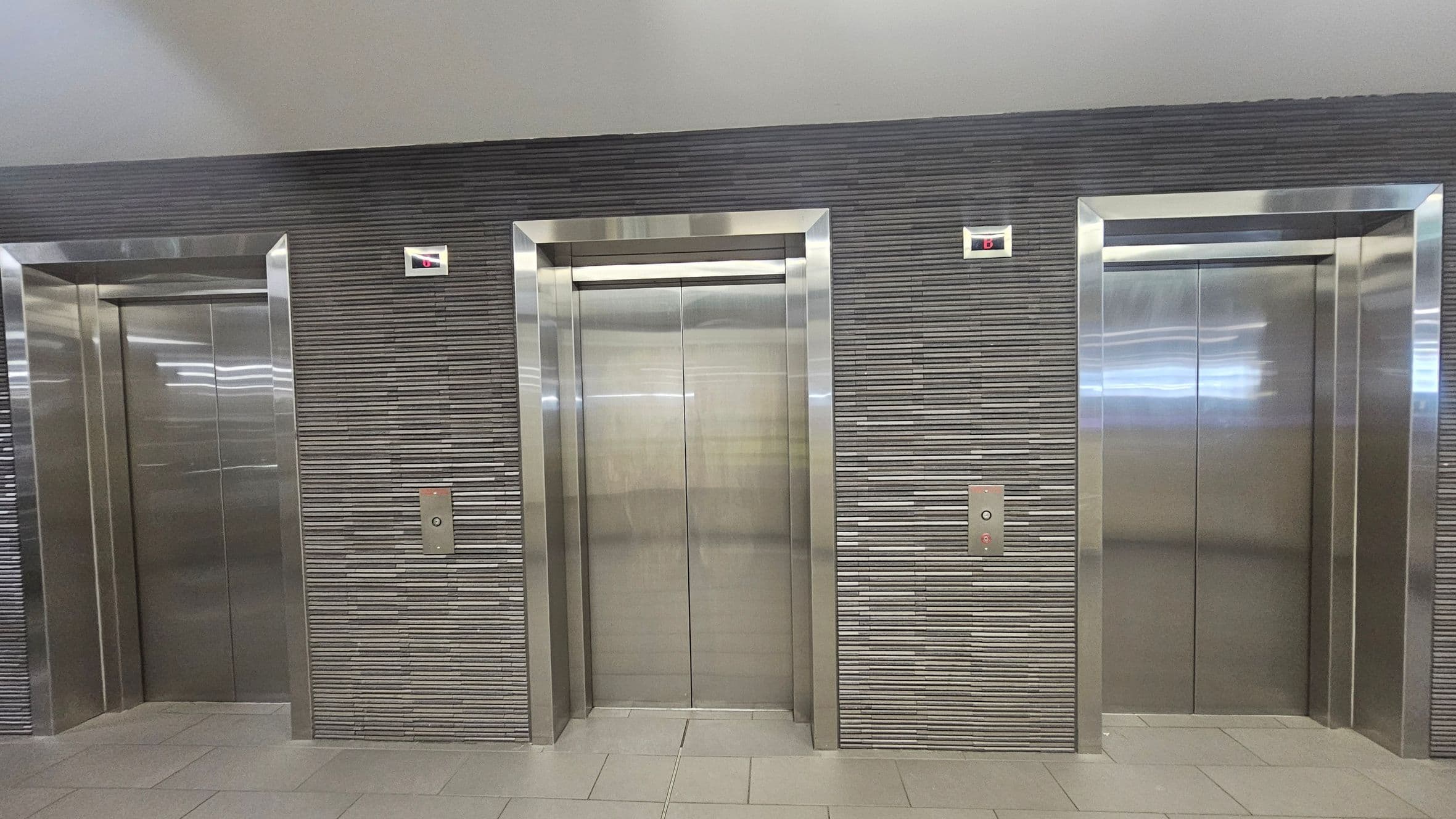 Three stainless steel lift doors are set into a wall with horizontal grey tiles. Each lift has a control panel with buttons and a digital display above showing the letter "B". The floor is tiled in a light grey colour, and the ceiling is smooth and light-coloured. The lifts are evenly spaced along the wall.