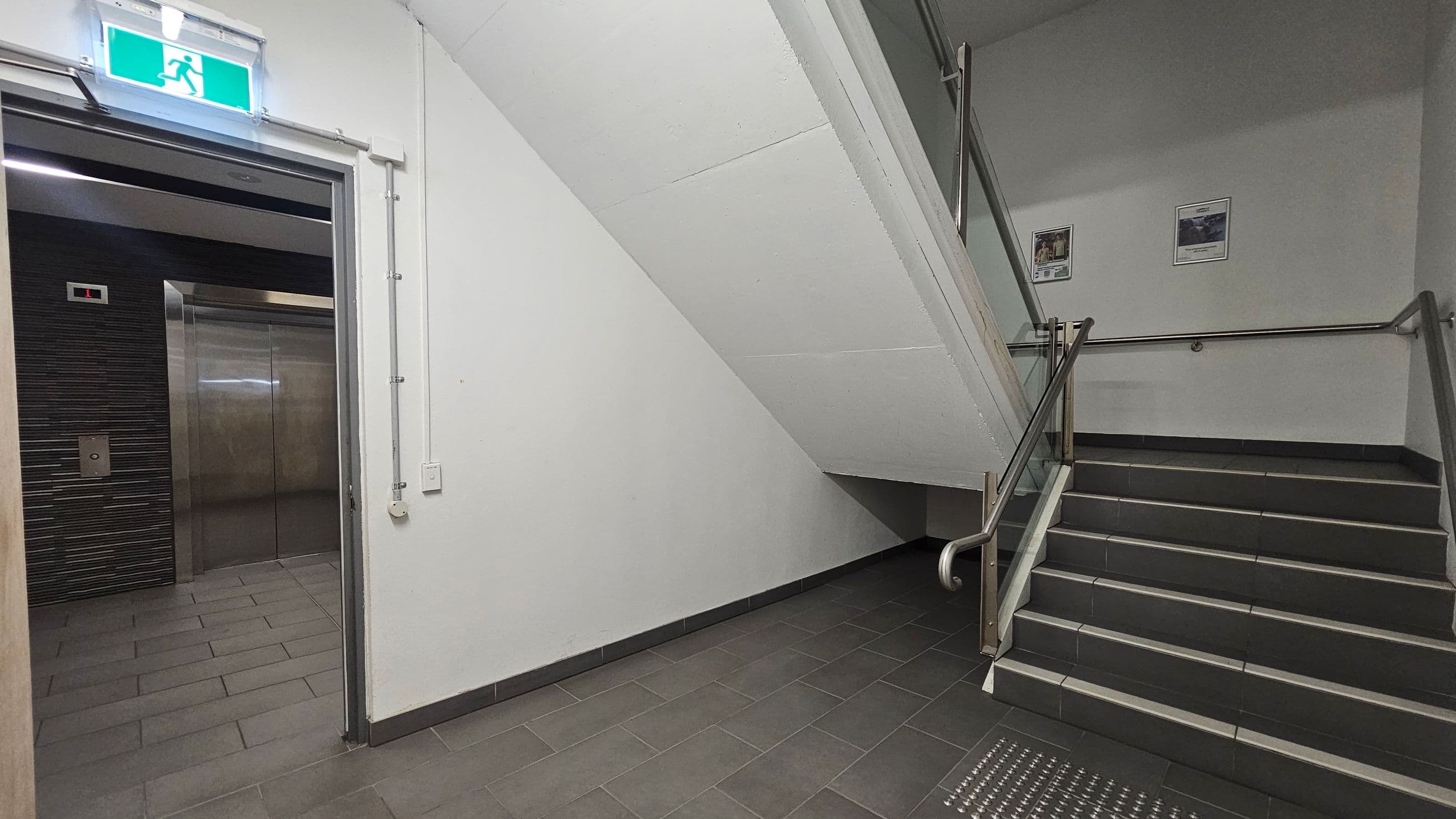 A corridor with a lift on the left, featuring metallic doors and a control panel. Above the lift is an illuminated green exit sign. On the right, a staircase with metal railings leads upwards. The walls are plain white, and two posters are mounted on the wall beside the stairs. The floor is covered with dark grey tiles, and the ceiling is white with visible piping.