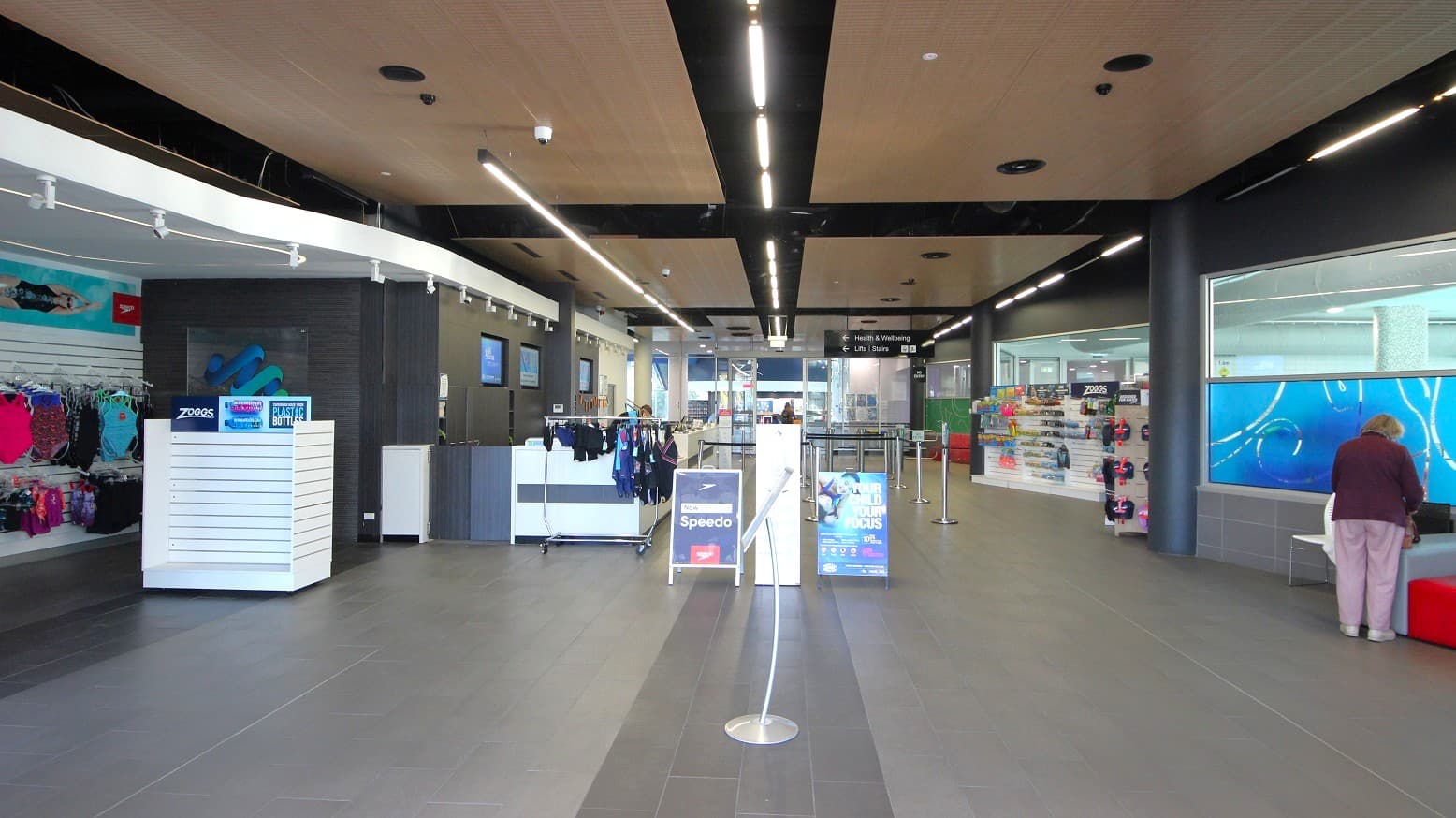 A spacious indoor area with a modern design. On the left, there are racks displaying swimwear and accessories. In the centre, a counter with hanging swim gear and promotional signs for Speedo. On the right, a person is standing near a bench, with a display of swimming goggles and accessories behind them. The floor is tiled in grey, and the ceiling features wooden panels with black beams and recessed lighting.