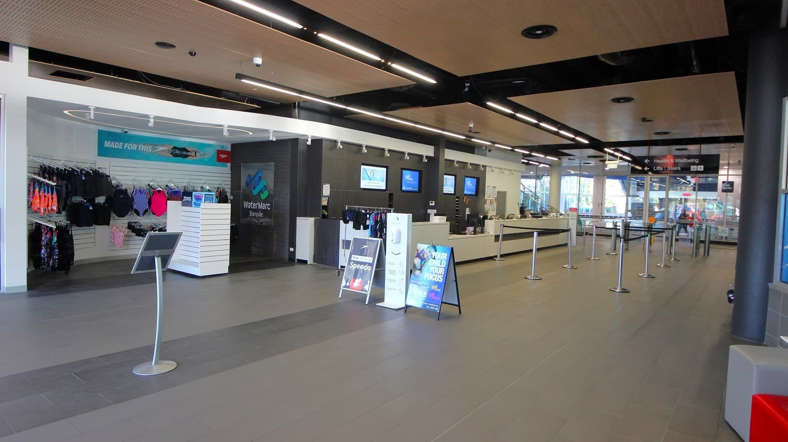 On the left, there are racks with swimwear and accessories against a white wall. In the centre, a stand with promotional signs is visible. To the right, a long reception counter with several monitors above it is present. The floor is a mix of grey and beige tiles, and the ceiling features wooden panels with black beams and recessed lighting. In the background, glass doors and windows allow natural light to enter the space.