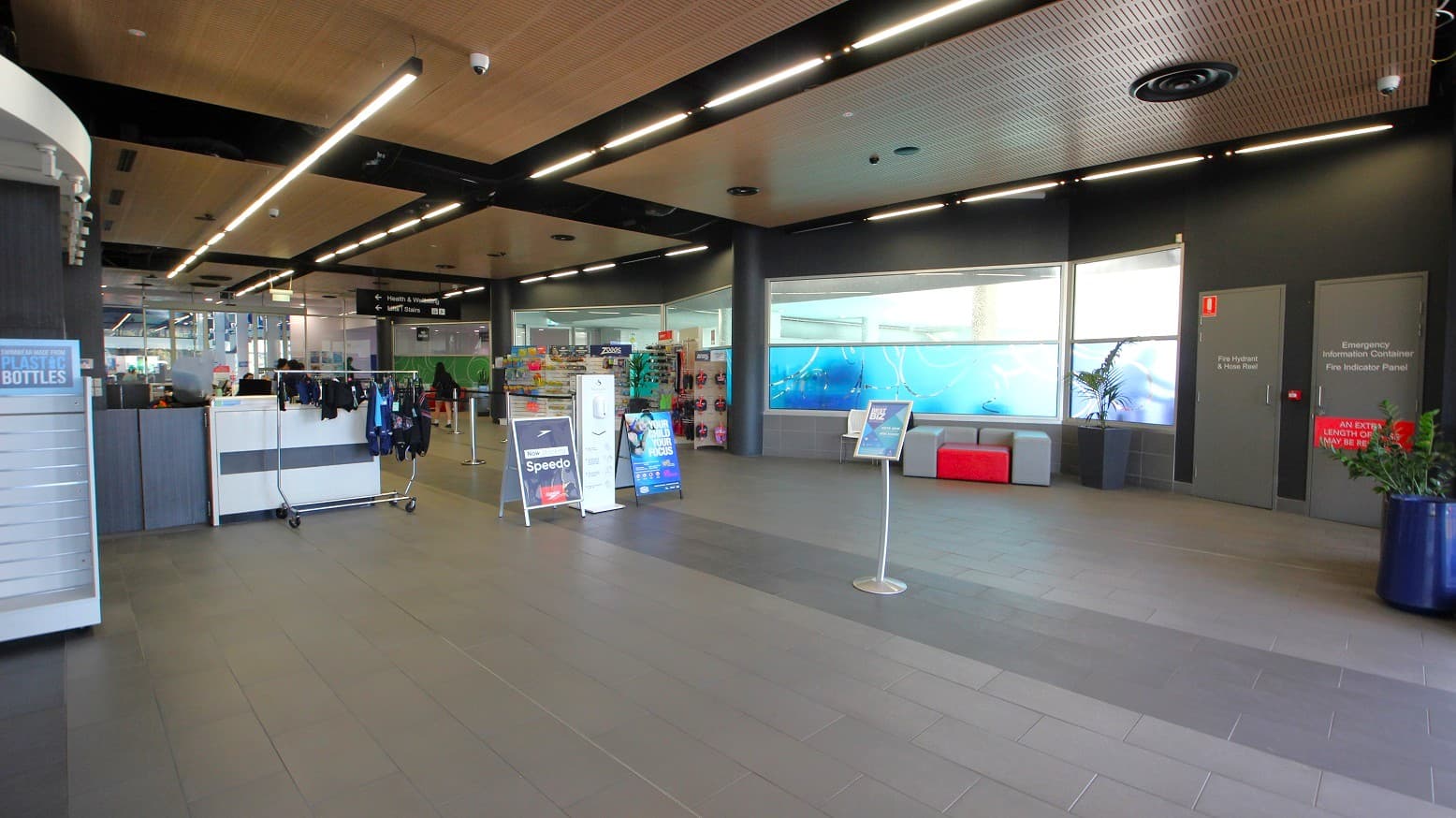 A spacious indoor area with a tiled floor and a ceiling featuring linear lights. On the left, there is a display with clothing items and a sign for Speedo. In the centre, there are promotional stands and a seating area with grey and red cushions. The right side has two grey doors with signage, and a large window with a blue aquatic-themed design. A potted plant is visible in the foreground on the right.
