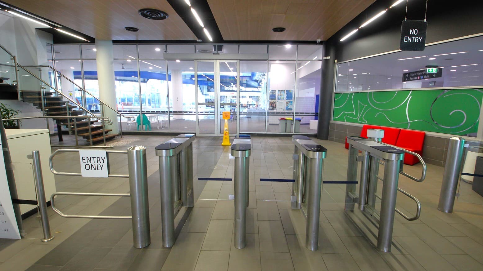 A modern indoor entrance area with turnstiles in the foreground, marked "ENTRY ONLY". To the left, a staircase with metal railings leads upwards. The background features glass doors and windows, revealing a brightly lit interior space. On the right, a green wall with abstract patterns and a red bench. The ceiling has recessed lighting and ventilation fixtures. A "NO ENTRY" sign hangs above the turnstiles. The floor is tiled in a neutral colour.