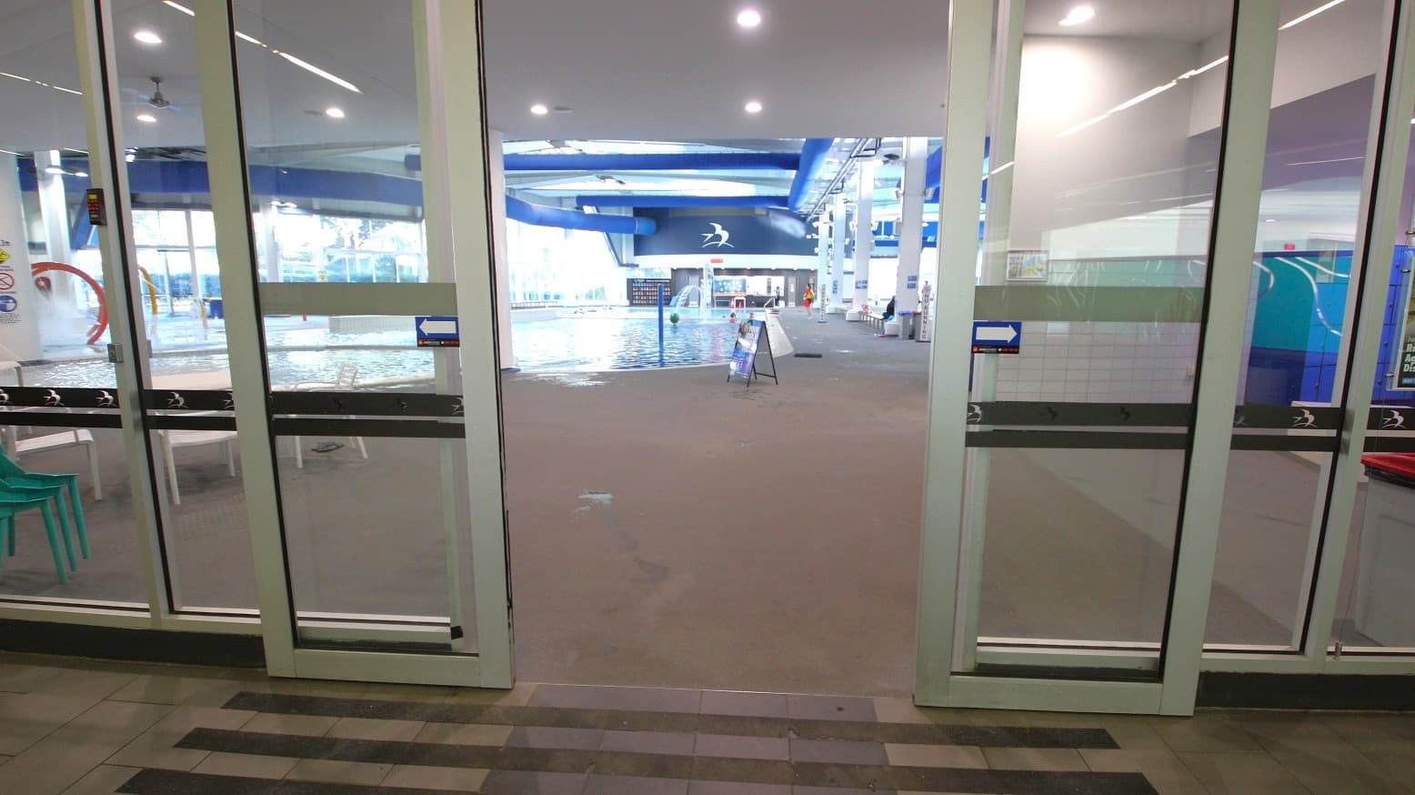 Glass doors open to an indoor swimming pool area. On the left, there are stacked green chairs and a glimpse of a play area with a red circular feature. In the centre, the pool is visible with a signboard on the poolside. The right side shows a blue and white wall with a design. The ceiling has recessed lighting and blue structural beams. The floor is tiled in a light brown colour.