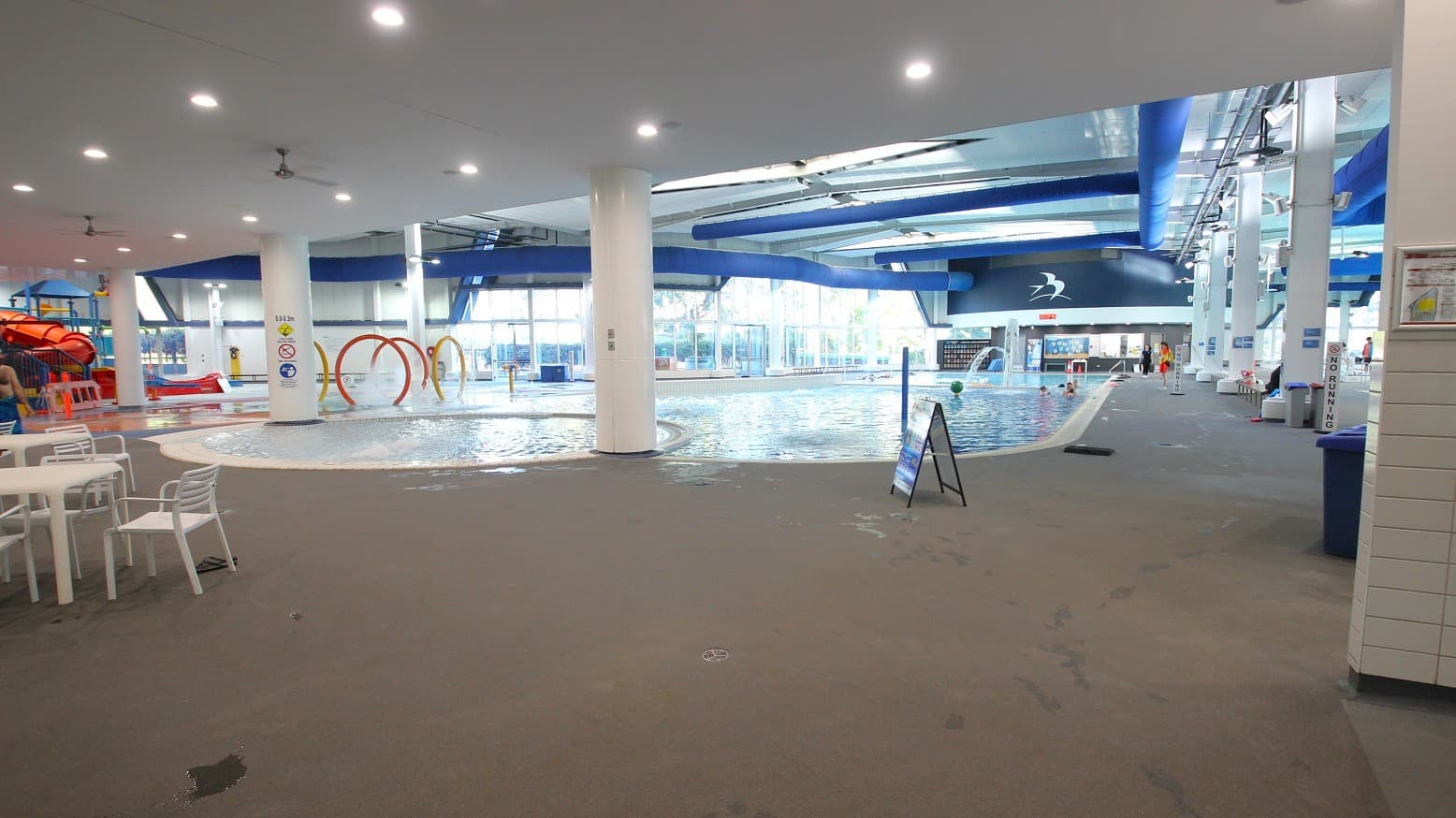 Indoor swimming pool area with a large pool in the centre. On the left, there are white tables and chairs, and a red water slide. In the background, large windows allow natural light to enter. The ceiling is white with blue ducts and recessed lighting. A signboard is placed near the pool, and there are various water play features like hoops. The floor is wet and made of non-slip material.