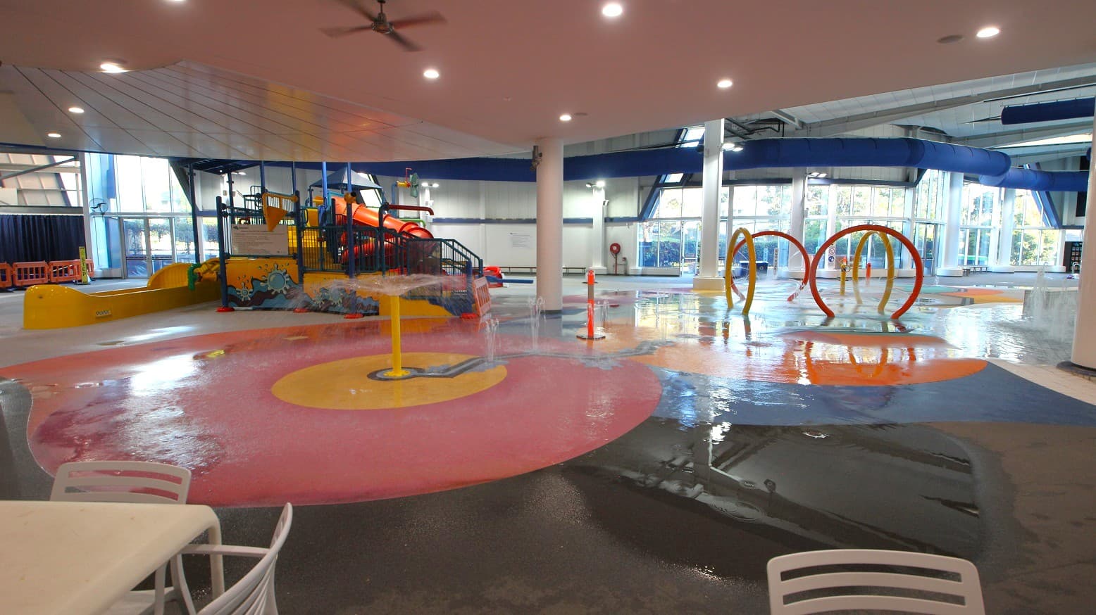 Indoor water play area with colourful features. On the left, a large play structure with slides in orange and yellow. In the centre, a pink and yellow splash pad with water jets. On the right, orange and yellow hoops for water play. The floor is wet and reflective, with a white table and chairs in the foreground. The ceiling is white with recessed lighting, and large windows in the background allow natural light in.