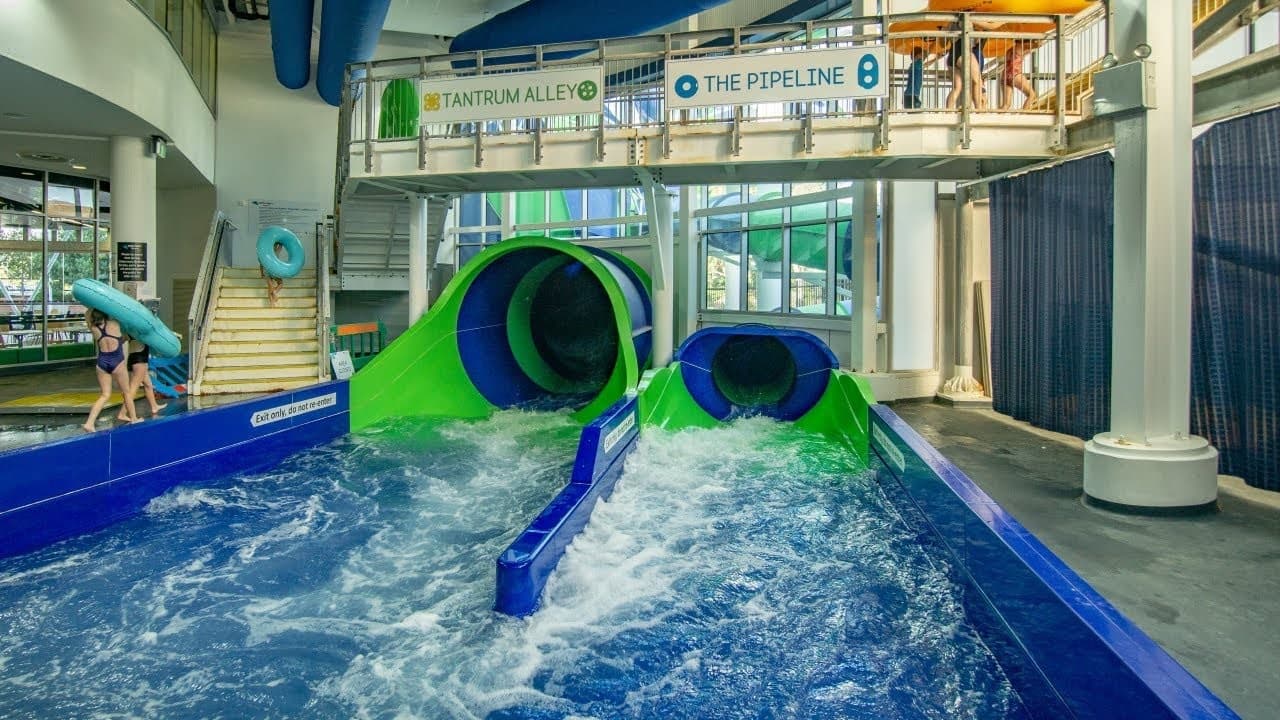 Indoor water park with two large water slides in the centre, one green and one blue. The slides empty into a pool with churning water. On the left, two people carry inflatable tubes up a staircase. Above the slides, signs read "Tantrum Alley" and "The Pipeline." The area is enclosed with large windows in the background, allowing natural light. The floor is wet and grey, and the ceiling has exposed blue pipes.