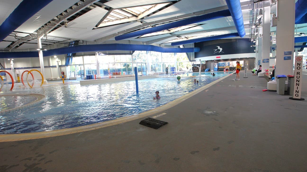 An indoor swimming pool area with a large pool in the centre. On the left, there are colourful hoops emerging from the water. The right side features a wide walkway with a "No Running" sign and several people sitting on benches. The ceiling is high with exposed beams and blue ducts. Large windows in the background allow natural light to enter, and a lifeguard stands near the pool. The floor is grey and textured.