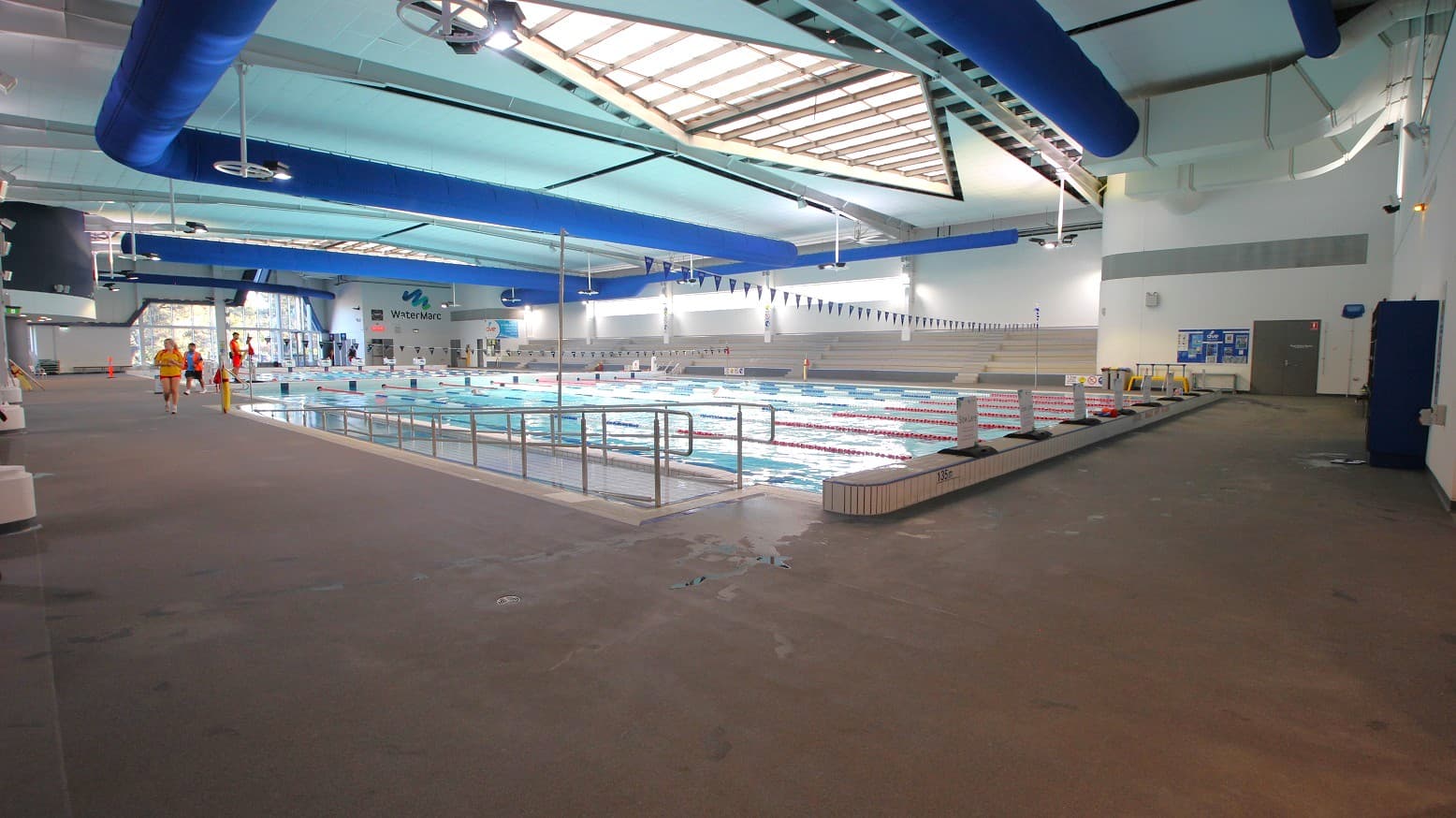 Indoor swimming pool with multiple lanes, surrounded by a wide deck area. On the left, a few people in swimwear and lifeguard attire walk along the deck. The pool is in the centre, with lane dividers and a railing. The ceiling features large blue ventilation ducts and skylights, allowing natural light. The walls are white with some signage, and there is a door on the right. The floor is a non-slip surface, and the area is well-lit.