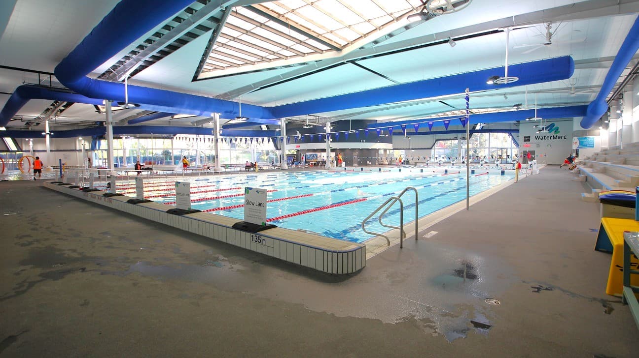 An indoor swimming pool with multiple lanes, separated by red and white lane dividers. On the left, there are lifeguards in orange vests near the pool's edge. The right side features a tiled pool deck with a metal handrail leading into the water. Blue ducts run along the ceiling, which has a large skylight. The floor is wet and textured for safety. Benches and a sign reading "WaterMarc" are visible in the background.