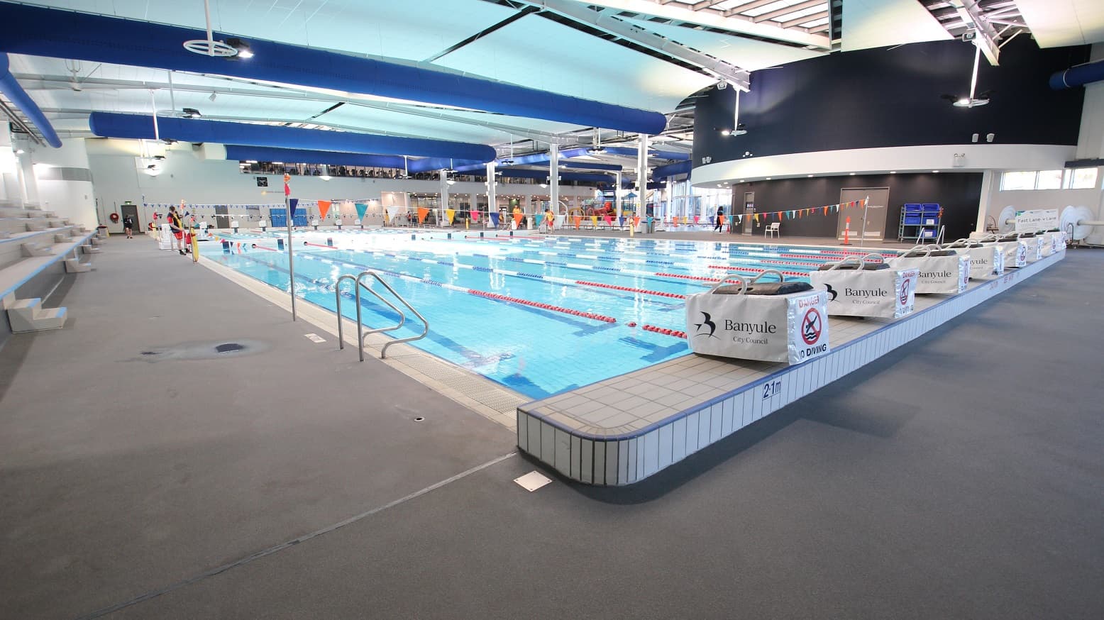 An indoor swimming pool with multiple lanes, separated by red lane dividers. On the left, there are tiered seating areas with grey benches. The pool deck is grey, with a metal handrail leading into the water. On the right, there are signs with "Banyule" and "No Diving" warnings. The ceiling features blue and white panels with exposed beams and lighting fixtures. In the background, there are people near the pool and a dark wall with windows above.