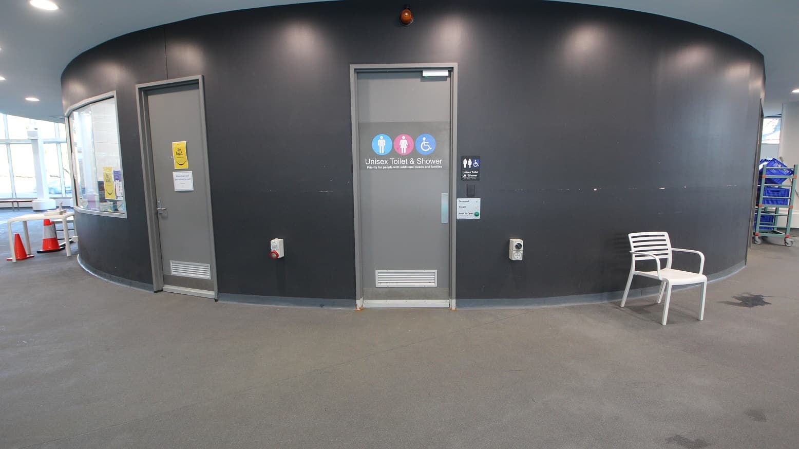 A curved, dark grey wall with two doors. The central door is marked "Unisex Toilet & Shower" with symbols for accessibility. To the left, another door has a yellow sign. A white chair is positioned on the right side of the image. The floor is carpeted in grey, and the ceiling is light-coloured with recessed lighting. On the far left, there are orange traffic cones and a window, while a blue trolley is partially visible on the right.