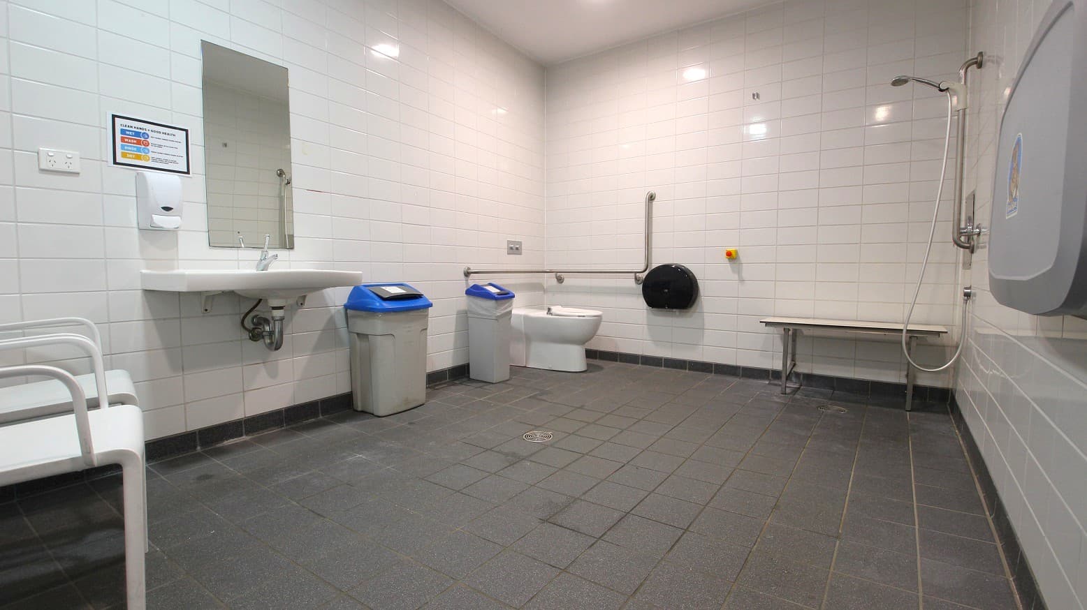 A spacious, accessible bathroom with white tiled walls and a dark tiled floor. On the left, there's a white sink with a mirror above it and a soap dispenser. Next to the sink are two bins with blue lids. In the centre, a toilet with a grab rail is visible. On the right, there's a shower area with a handheld showerhead and a foldable bench. A baby changing station is mounted on the wall. The ceiling is plain with recessed lighting.