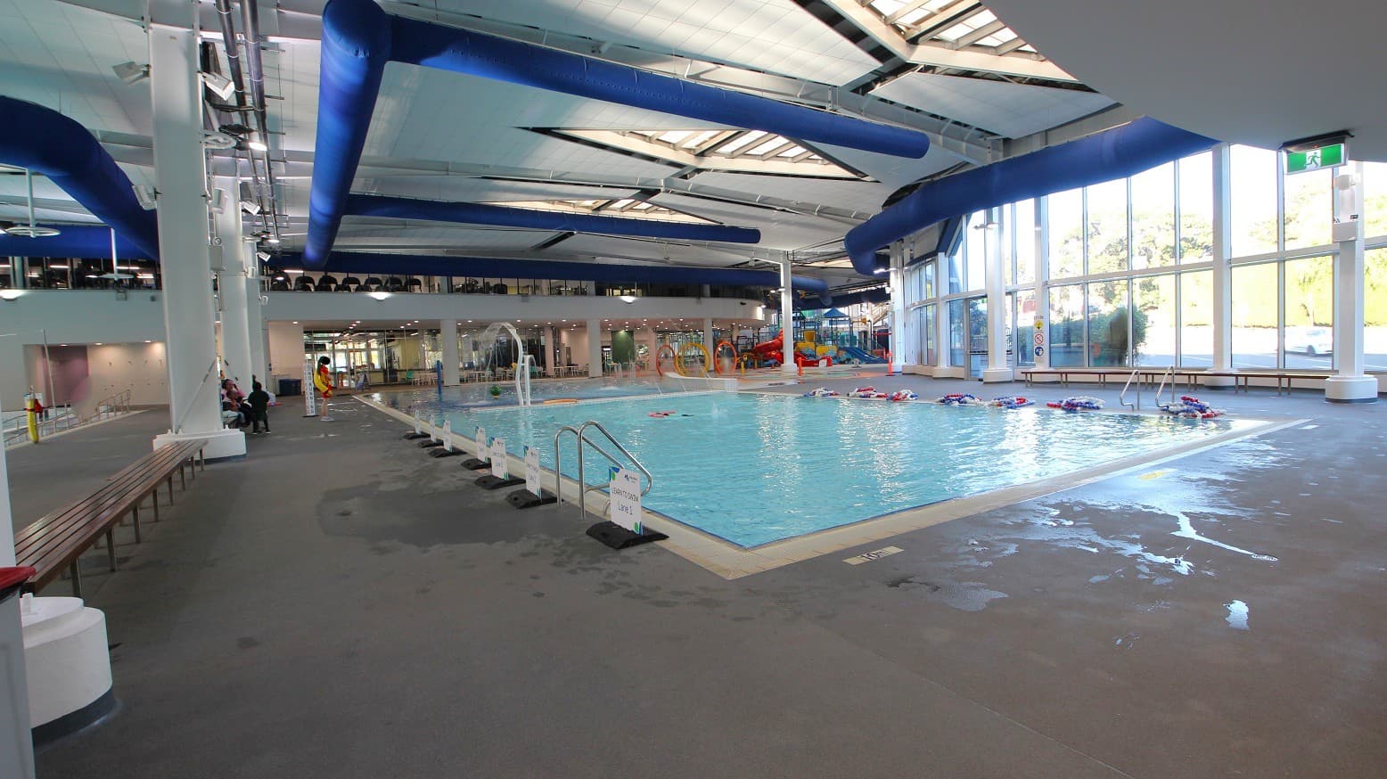 An indoor swimming pool area with a large pool on the right, surrounded by a tiled deck. Several flotation devices are floating in the water. On the left, there are benches and a few people seated. The ceiling features blue ventilation ducts and skylights, allowing natural light to illuminate the space. Large windows on the right wall provide additional light and a view outside. The floor is wet in places, indicating recent use.