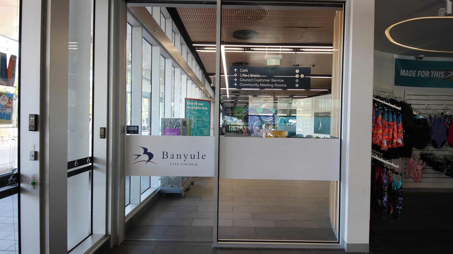 Glass entrance doors with a sign reading "Banyule City Council" in the centre. To the left, a keypad and glass panels. Inside, a directory sign points to a café, lifts, stairs, council customer service, and community meeting rooms. The interior has a wooden ceiling and visible lighting. On the right, a retail area displays colourful clothing on racks.