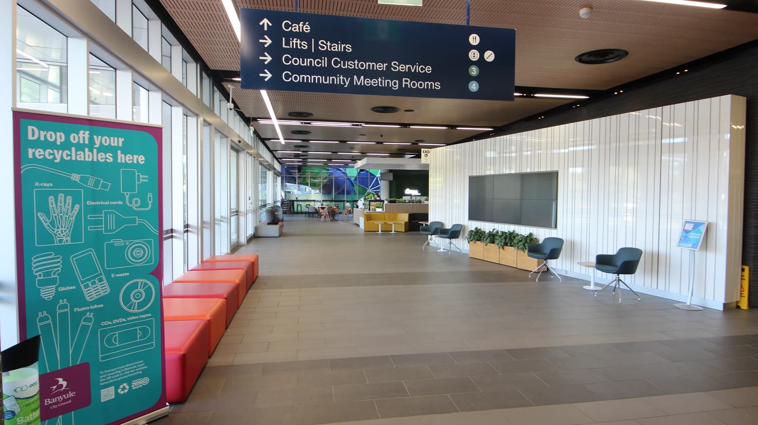 A spacious indoor corridor with a high ceiling featuring linear lights. On the left, large windows allow natural light to flood in, with a teal sign for recyclables and red seating along the wall. On the right, a white panelled wall with a mounted screen, flanked by two chairs and a row of potted plants. Overhead, a blue sign directs to a café, lifts, council customer service, and meeting rooms. In the background, a café area with tables and chairs is visible.