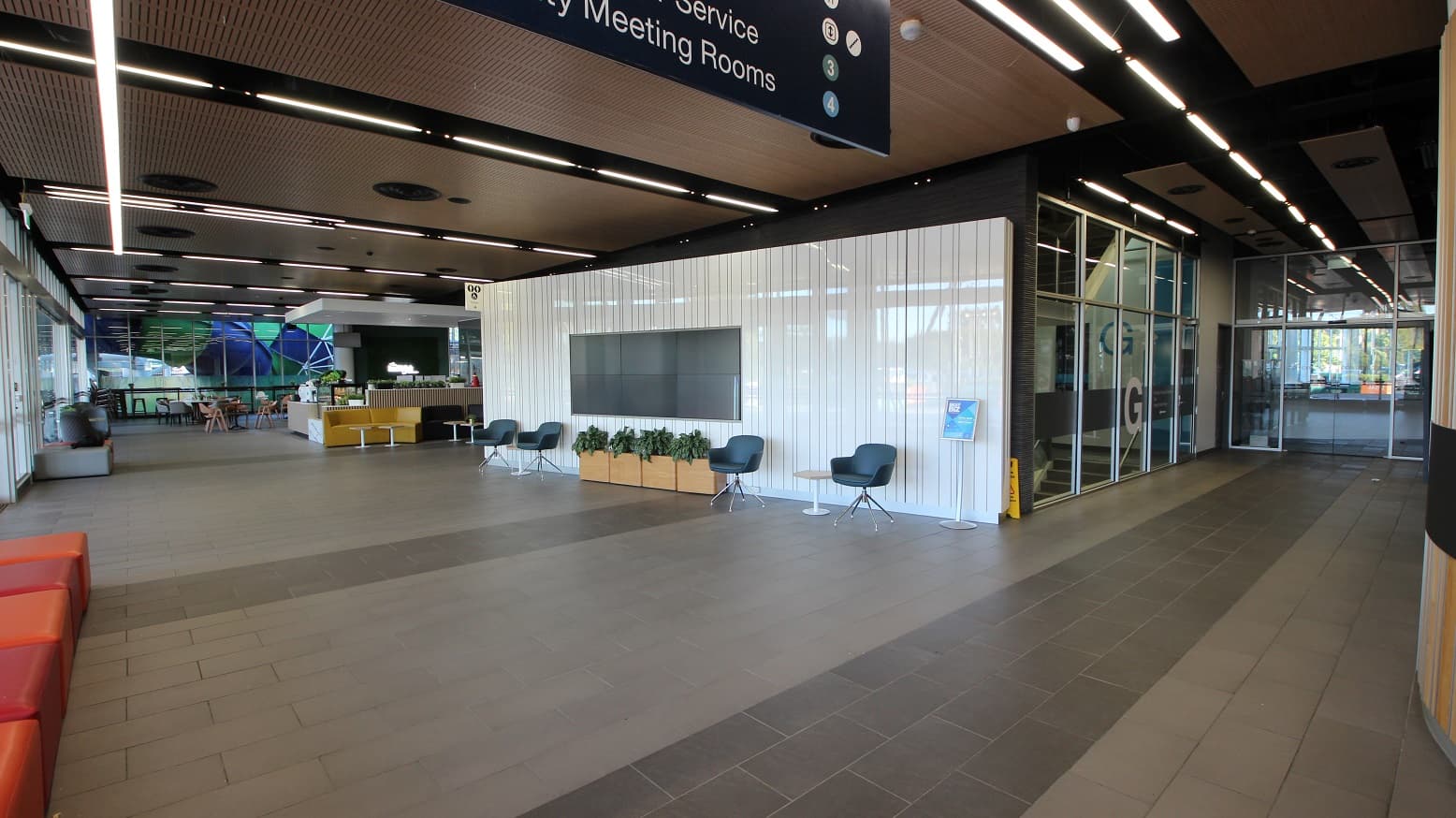 A modern interior space with a high ceiling featuring linear lights. On the right, there's a glass entrance with a sign above. In the centre, a white wall with a large screen is flanked by chairs and potted plants. On the left, a seating area with orange couches and a reception desk is visible. The floor is tiled in shades of grey, and the ceiling has a wood panel design with circular vents.