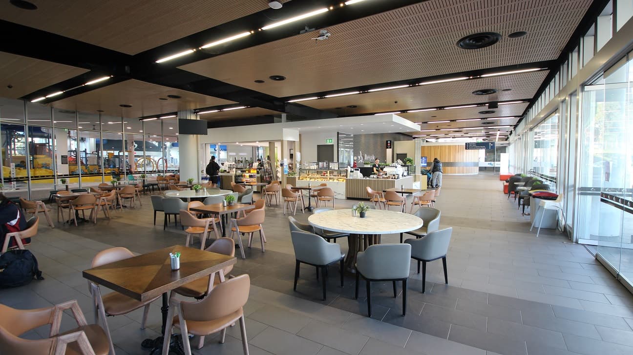 A spacious, modern café area with a variety of seating options, including round and square tables surrounded by wooden chairs. The left side features a glass wall with a view of an escalator and some equipment. In the centre, there's a counter with food displays and a person standing nearby. The right side has large windows allowing natural light to fill the space, with additional seating along the window. The ceiling has a wooden finish with black beams and recessed lighting.