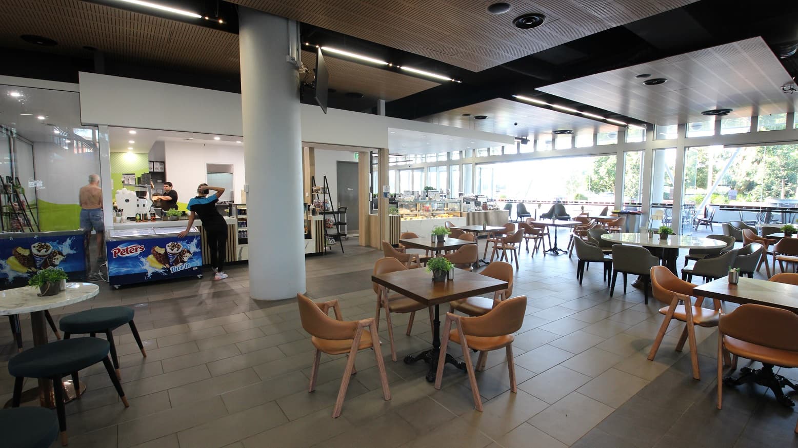 A modern café interior with a spacious seating area. On the left, there is a counter with ice cream freezers branded "Peters" and staff behind the counter. In the middle, several wooden tables and chairs are arranged neatly, each with a small plant as a centrepiece. The right side features large windows allowing natural light to flood in, with more seating visible outside. The ceiling has a mix of wooden and black panels, and the floor is tiled.