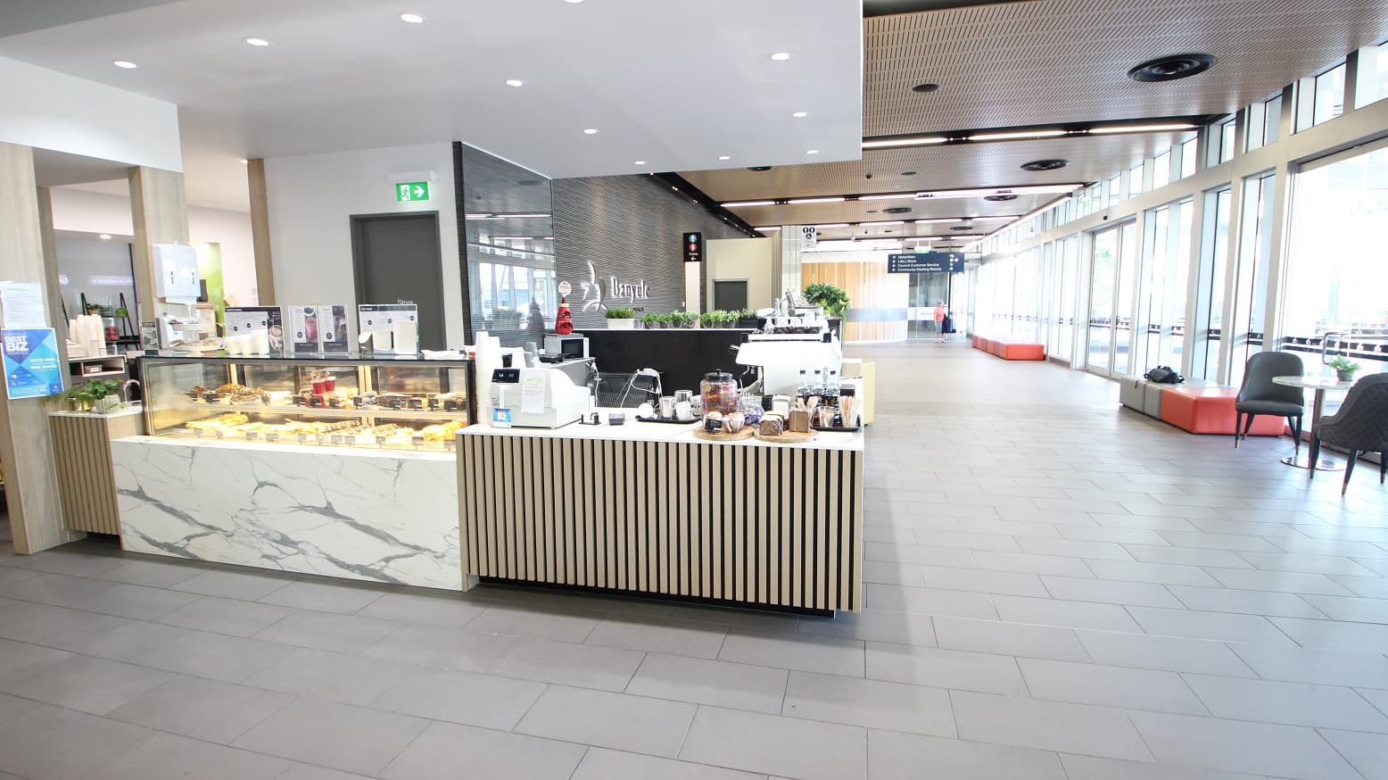 A modern café interior with a marble and wood counter on the left displaying pastries and a coffee machine. The floor is tiled in grey, and the ceiling has recessed lighting. On the right, large windows allow natural light to flood in, with seating areas featuring grey chairs and orange benches. In the background, a corridor leads to other areas, with signs and more seating visible.